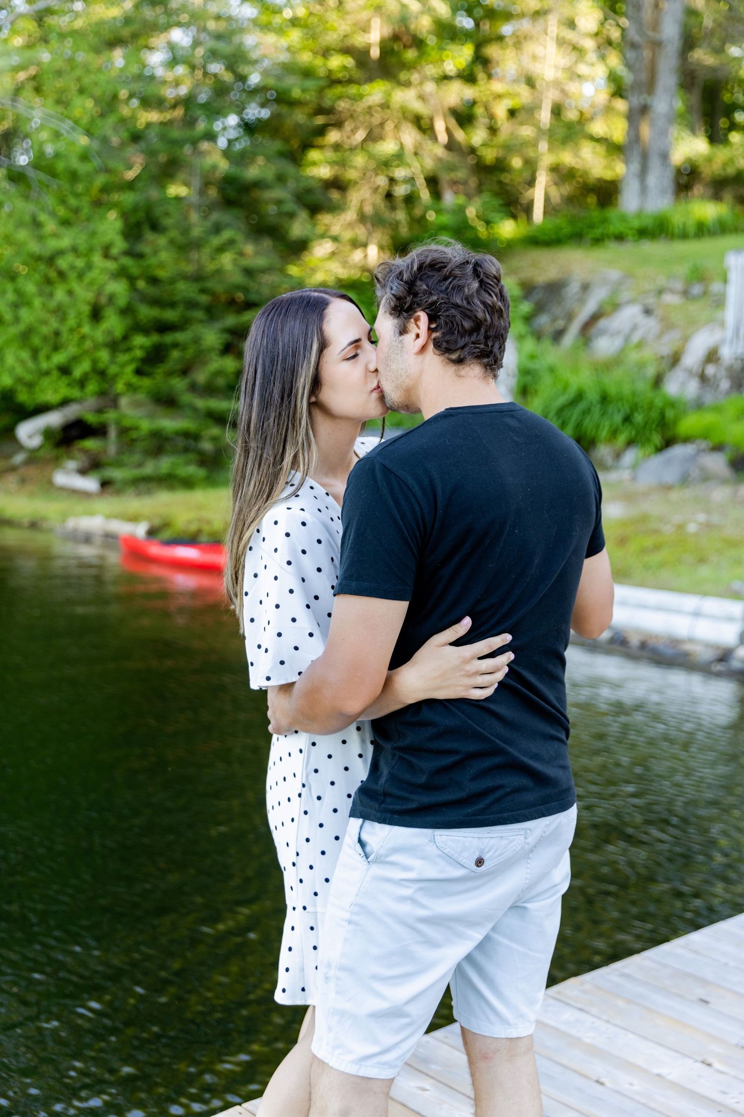 A couple shares a kiss on a wooden dock with a serene lake and red kayak in the background.