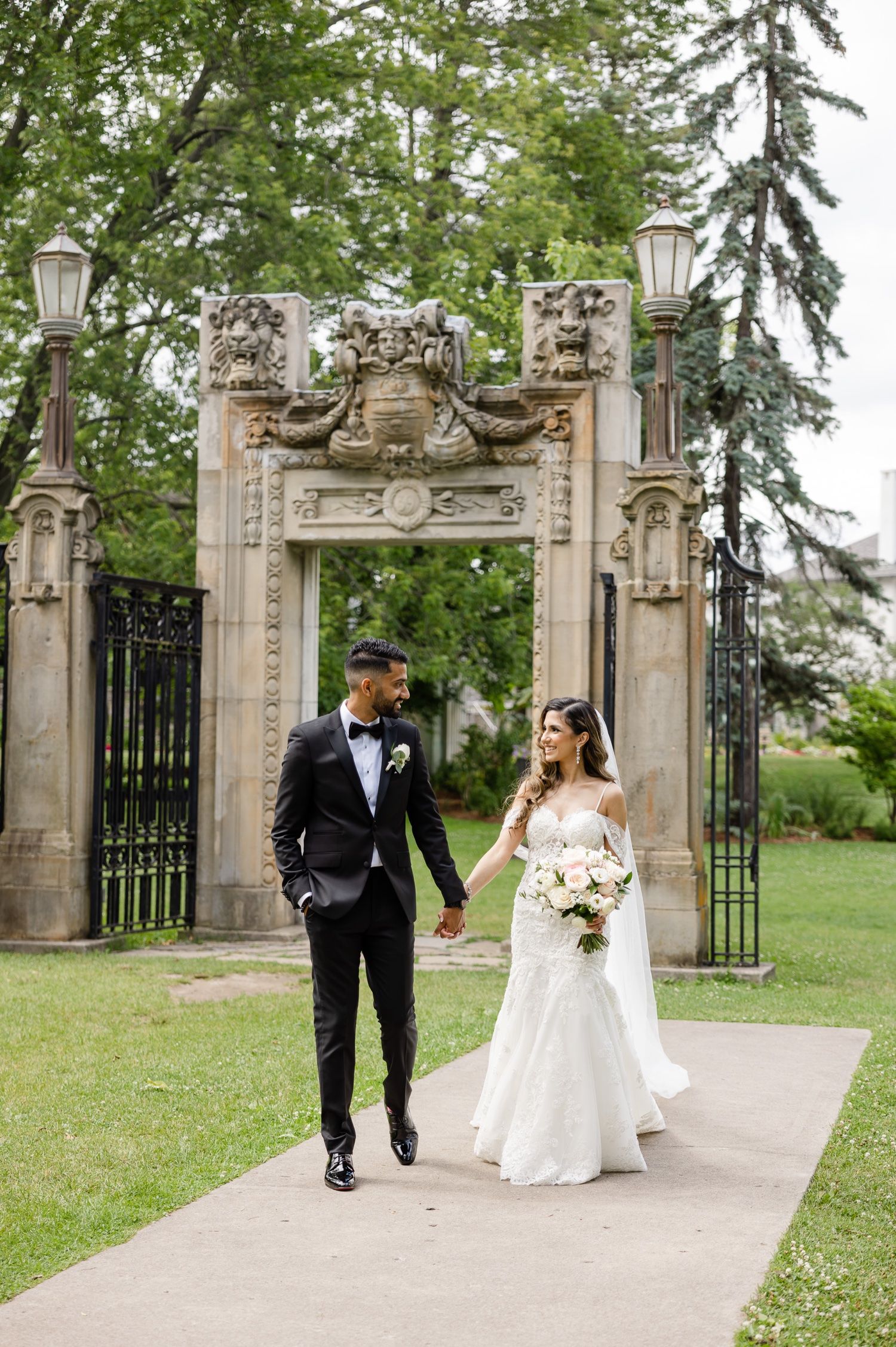 Wedding couple walking through an elegant stone archway entrance surrounded by lush greenery.