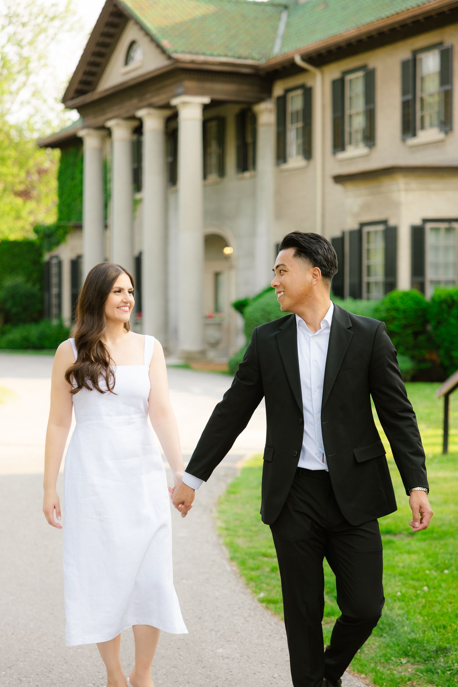 Couple shares joyful moment while walking hand in hand by mansion with columns.