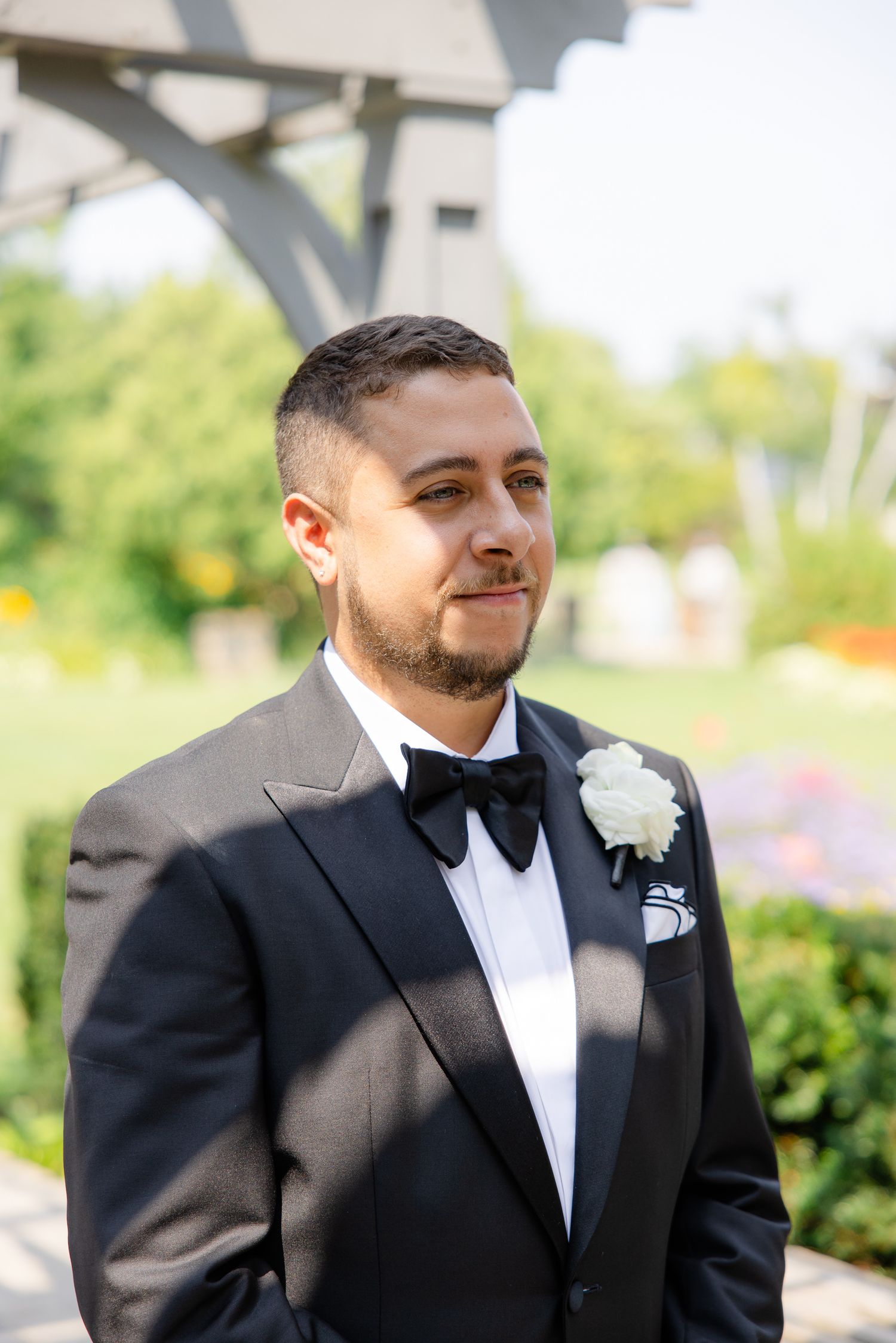 Groom in classic black tuxedo and bowtie with boutonniere stands in garden setting.