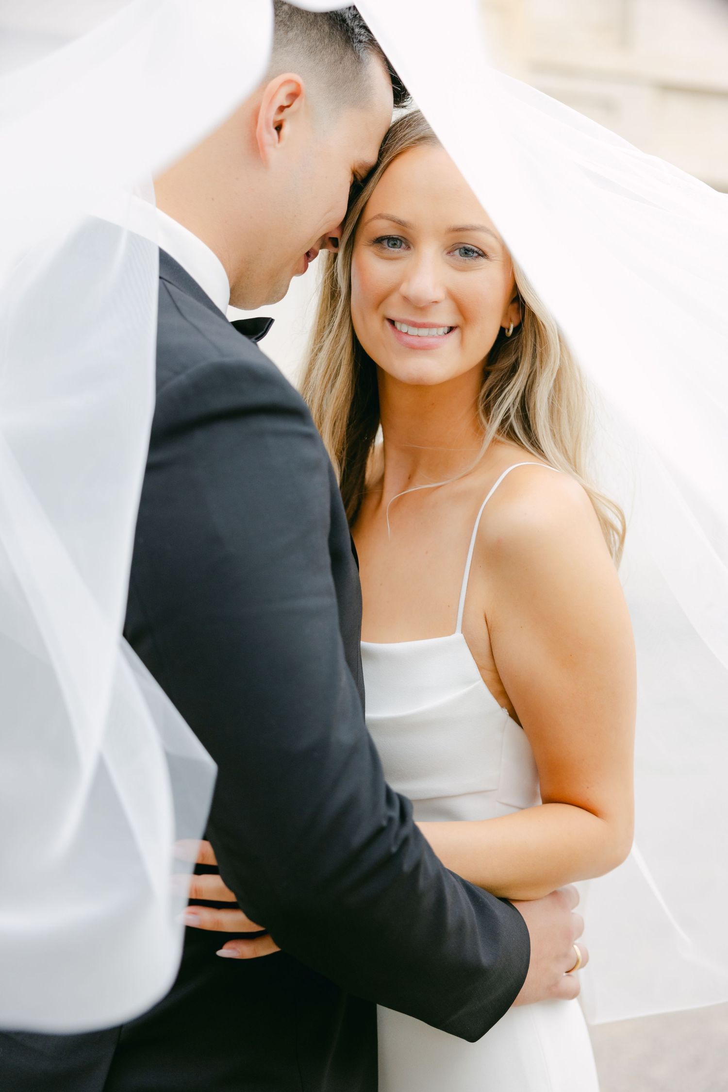 Romantic wedding portrait under veil with bride in sleek dress and groom in classic tuxedo.