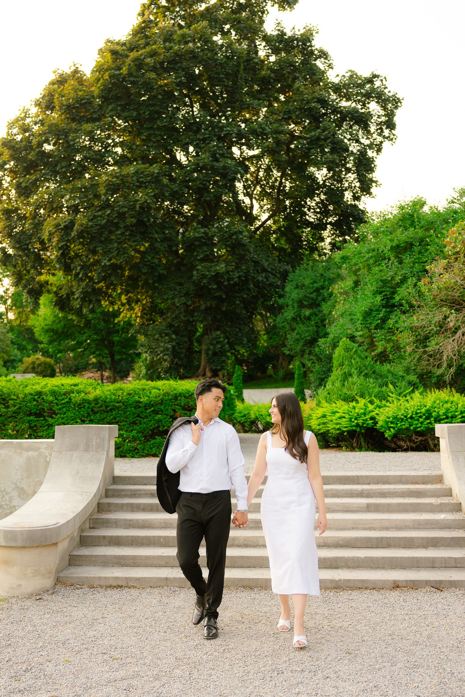 Couple walks up elegant stone stairs surrounded by manicured hedges and mature trees in a formal garden.