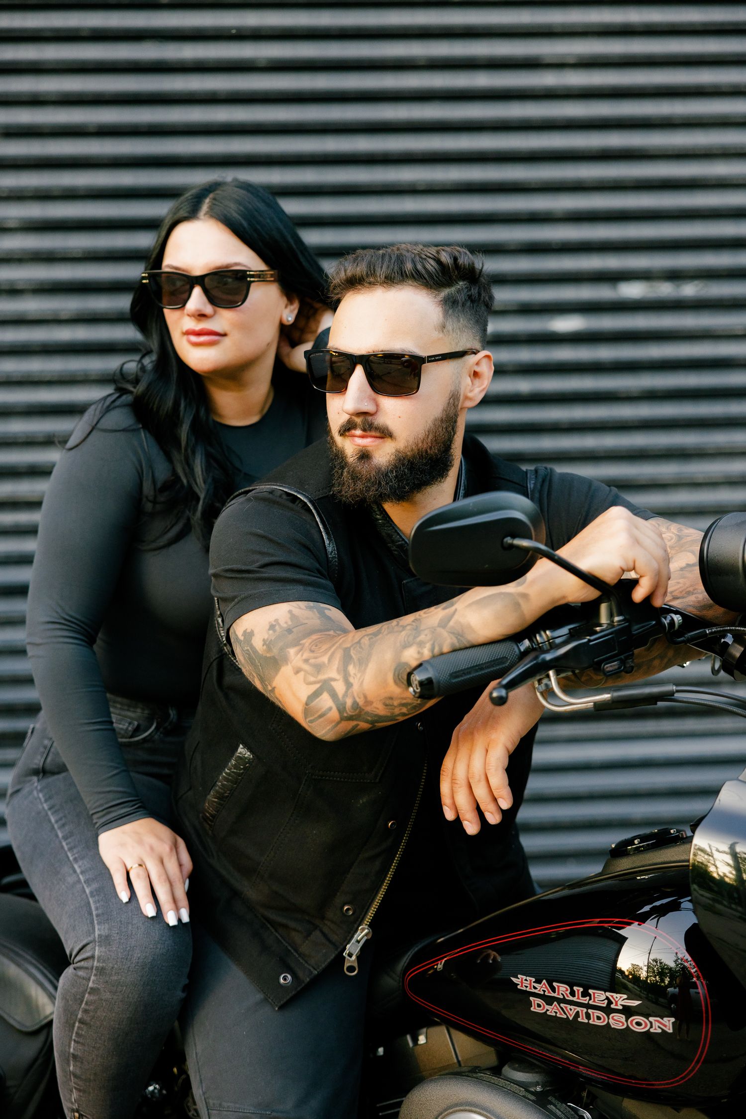 A fashionable couple in sunglasses poses with their Harley Davidson motorcycle against metal siding.