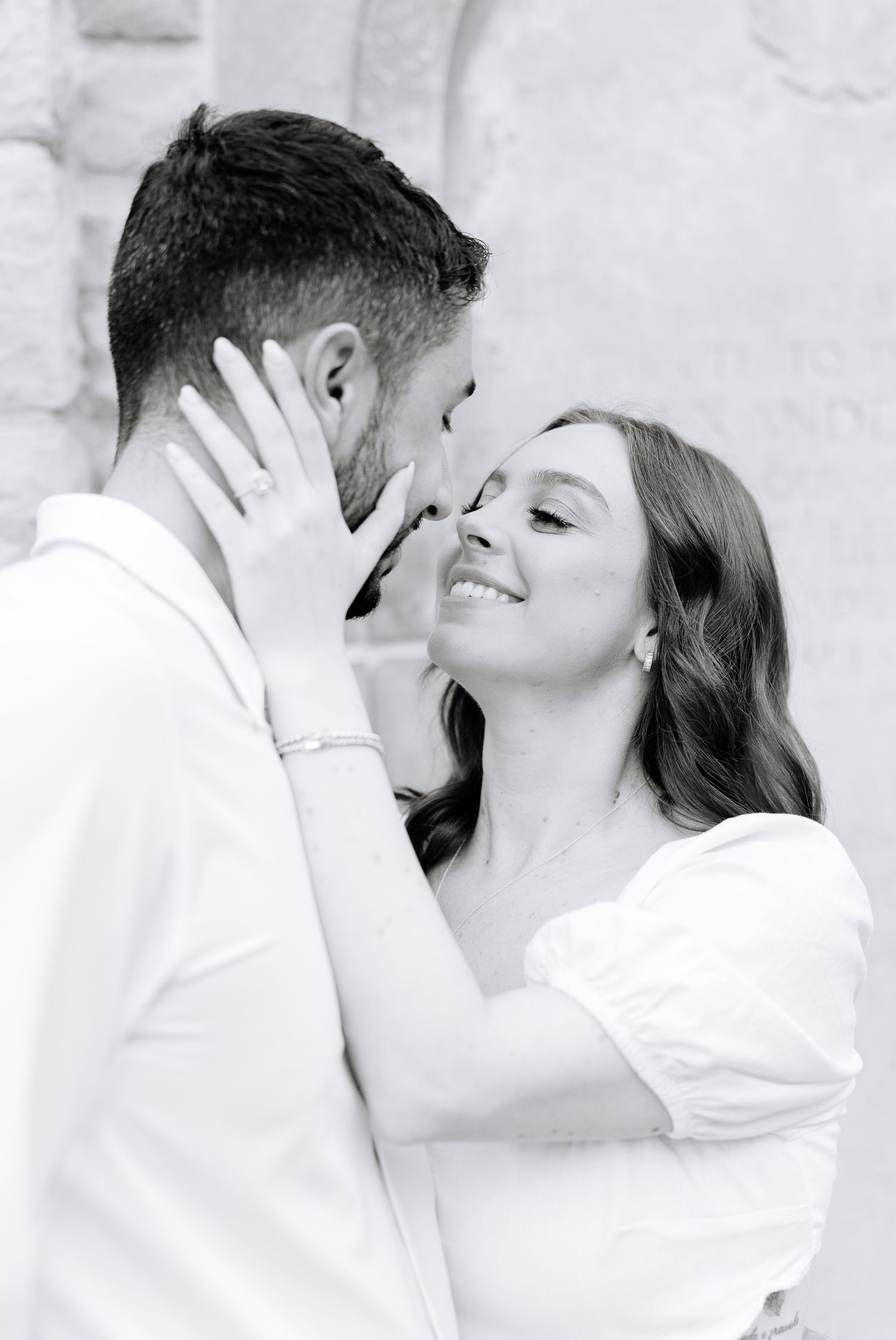 Black and white close-up of tender moment between two people against stone wall.