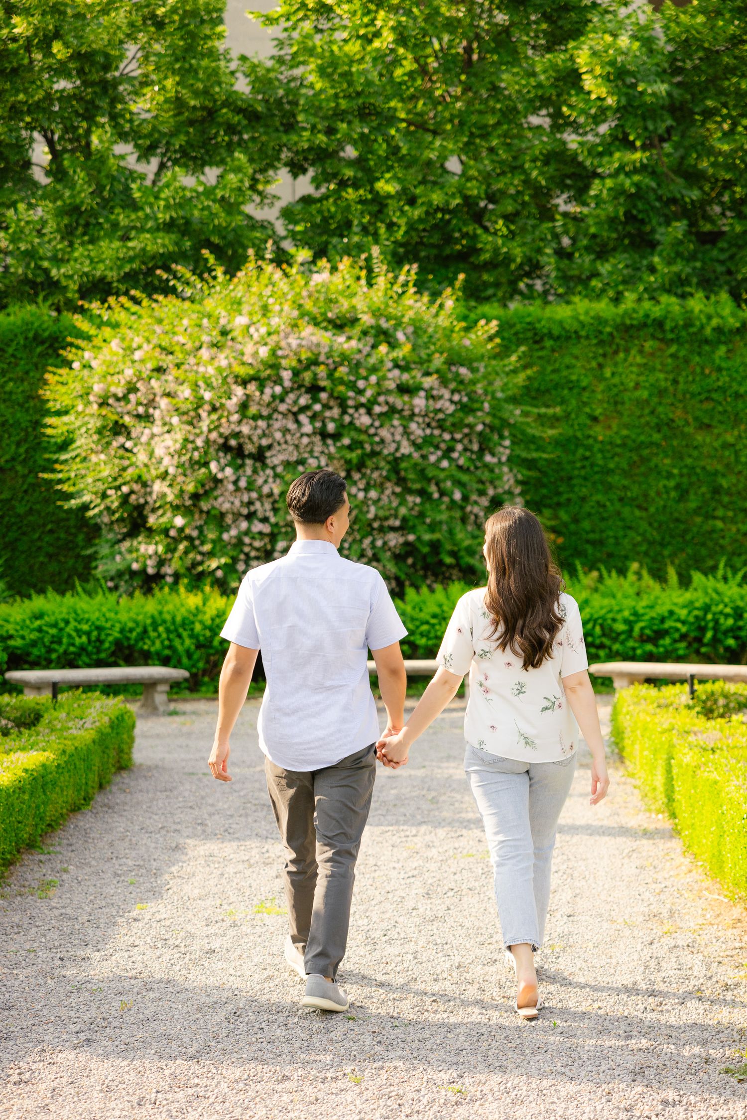A couple walks hand in hand down a manicured garden path surrounded by blooming flowers and hedges.