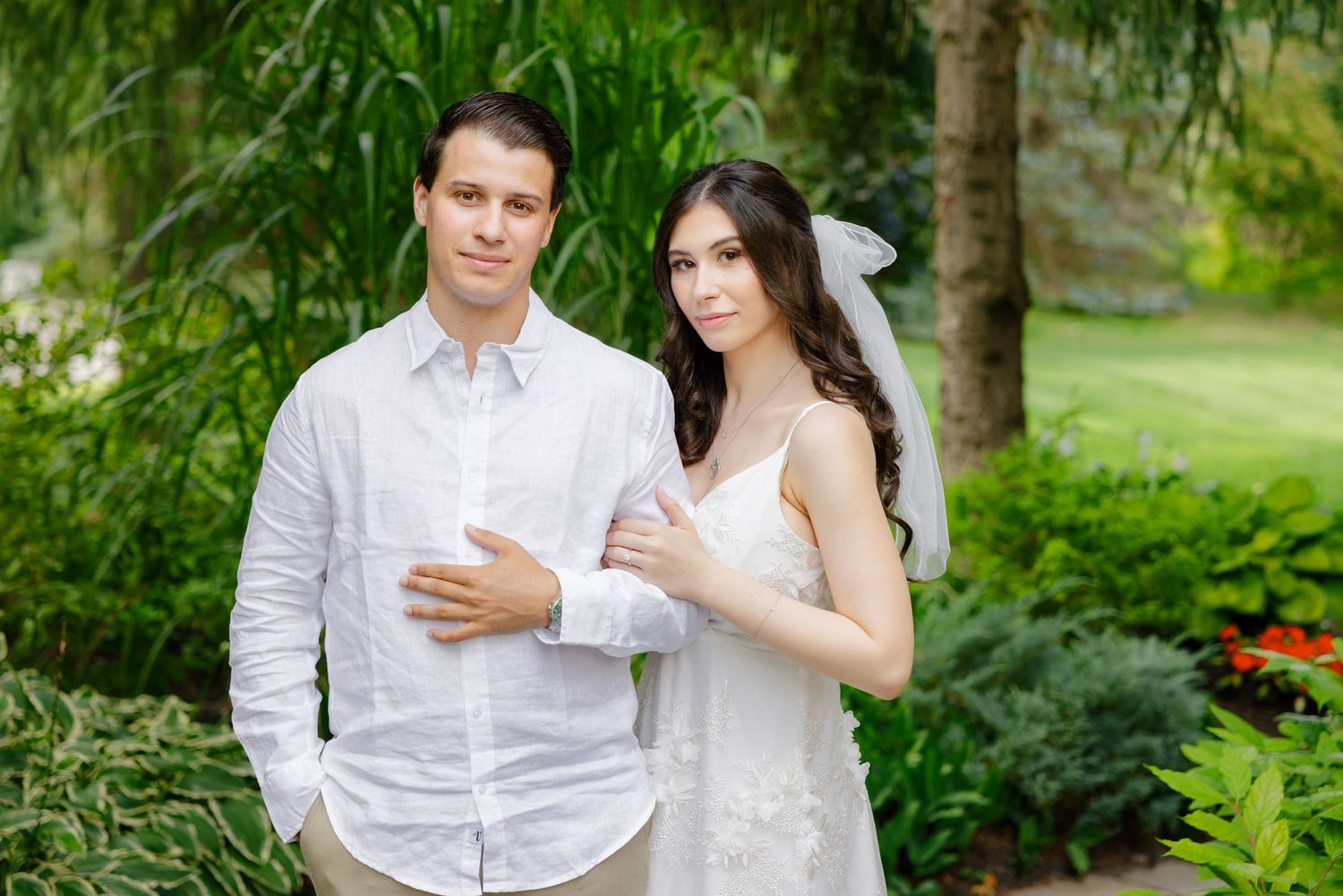 A couple in white stands together in a beautifully landscaped garden setting.