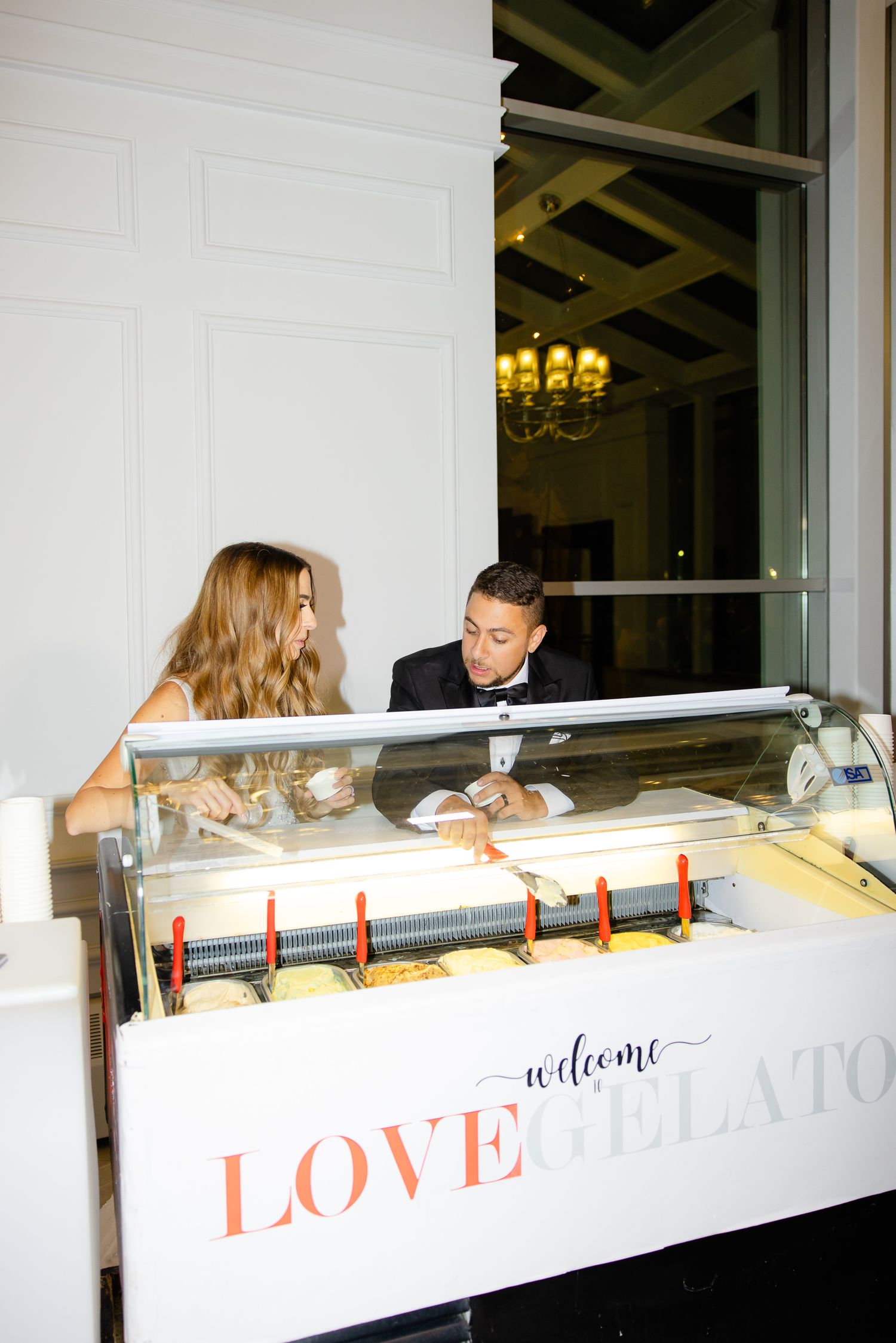 Couple enjoying gelato from a white ice cream display cart with 'LOVE welcome' text at an elegant event.