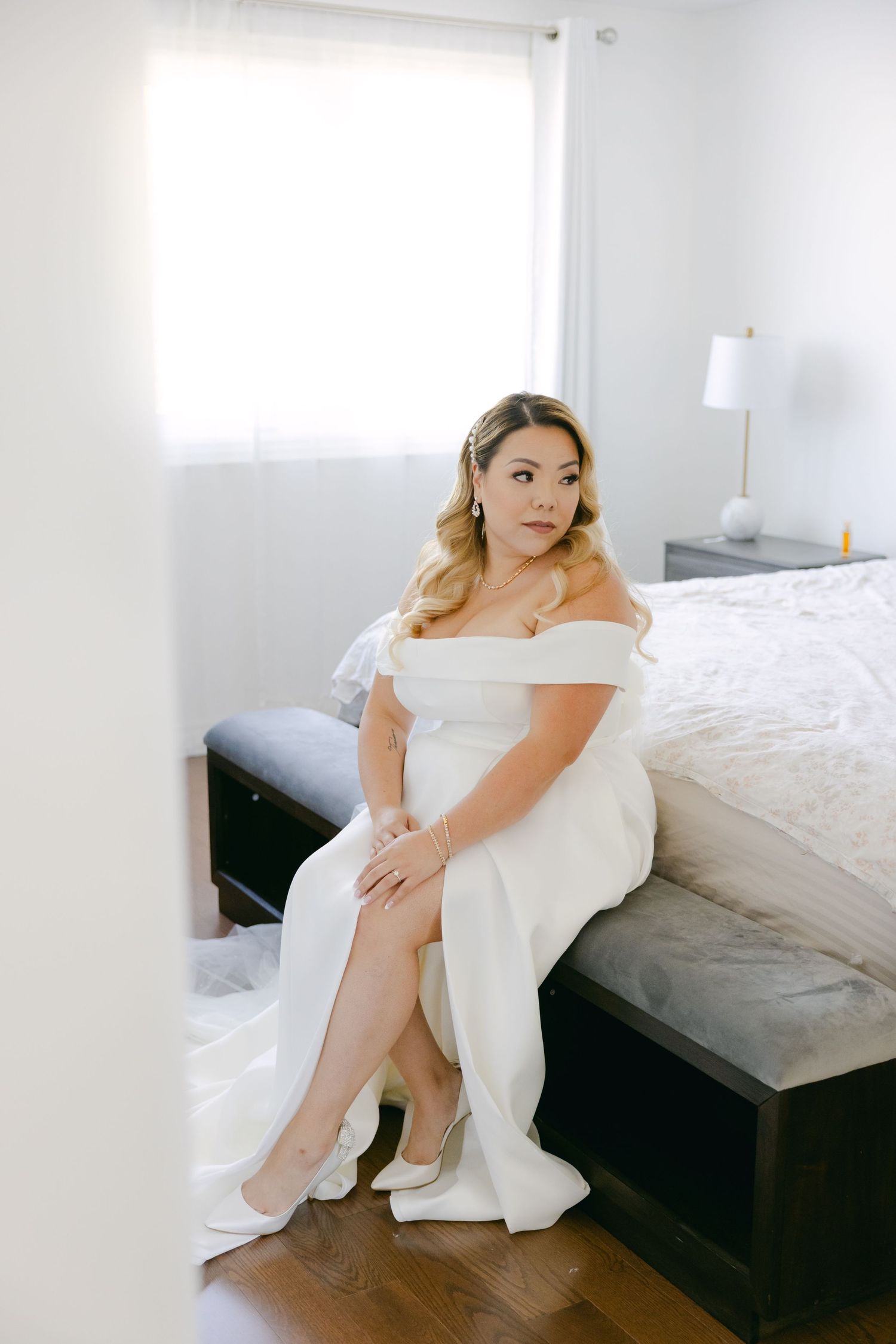 A bride in a classic white off-shoulder gown sits elegantly on the edge of a bed in a bright room.