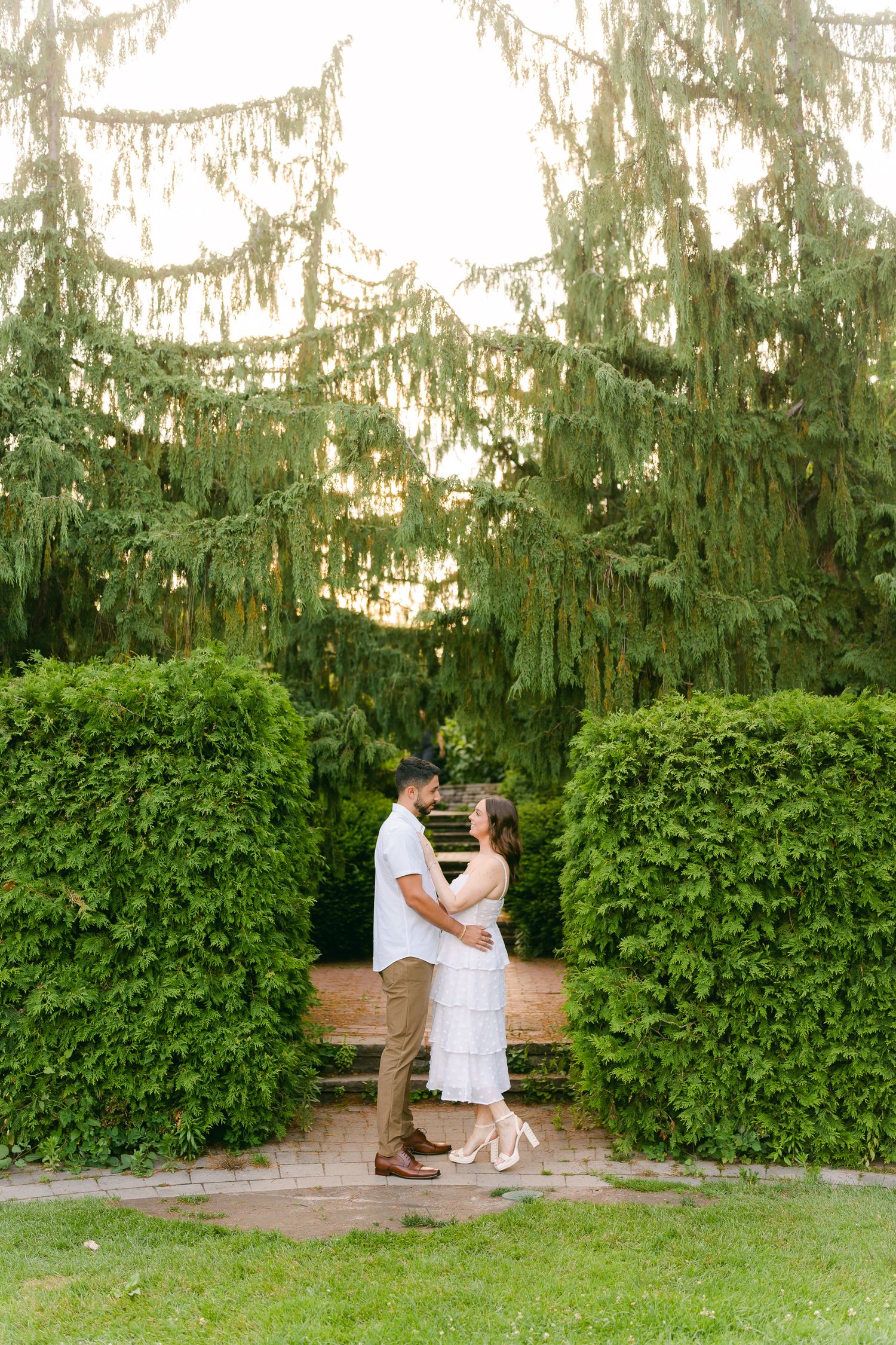 A romantic couple embraces in a garden pathway framed by lush hedges and weeping willow trees at sunset.