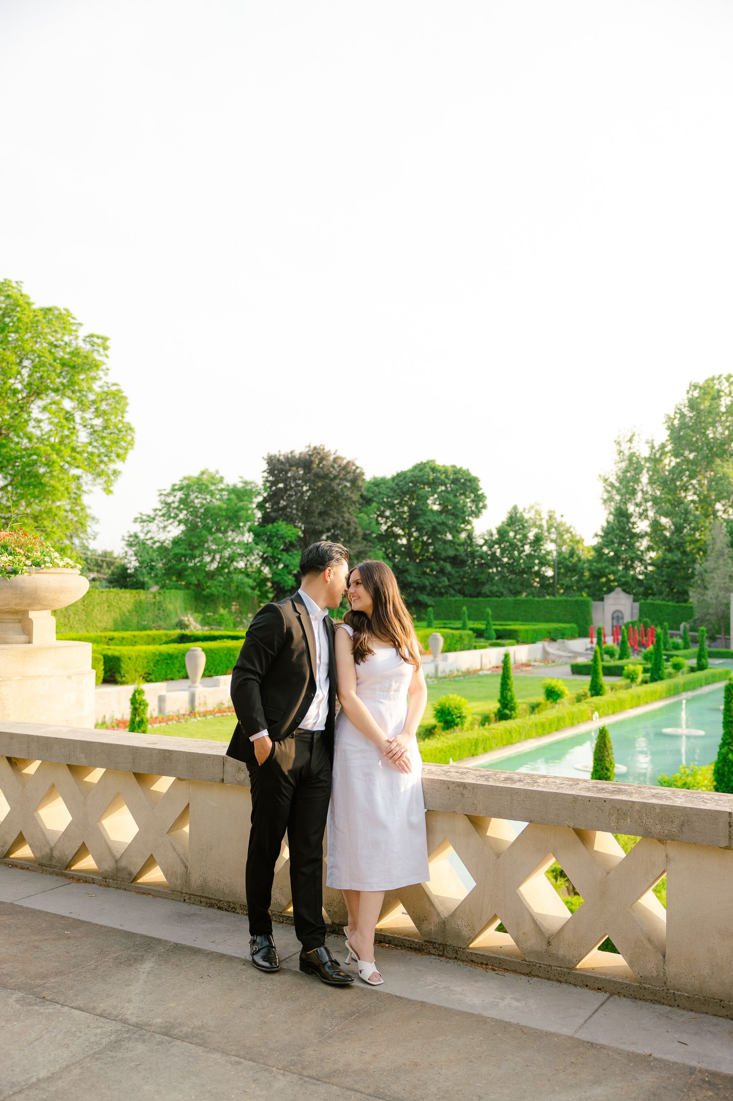 A romantic embrace on a stone terrace with manicured gardens and water features in the background.
