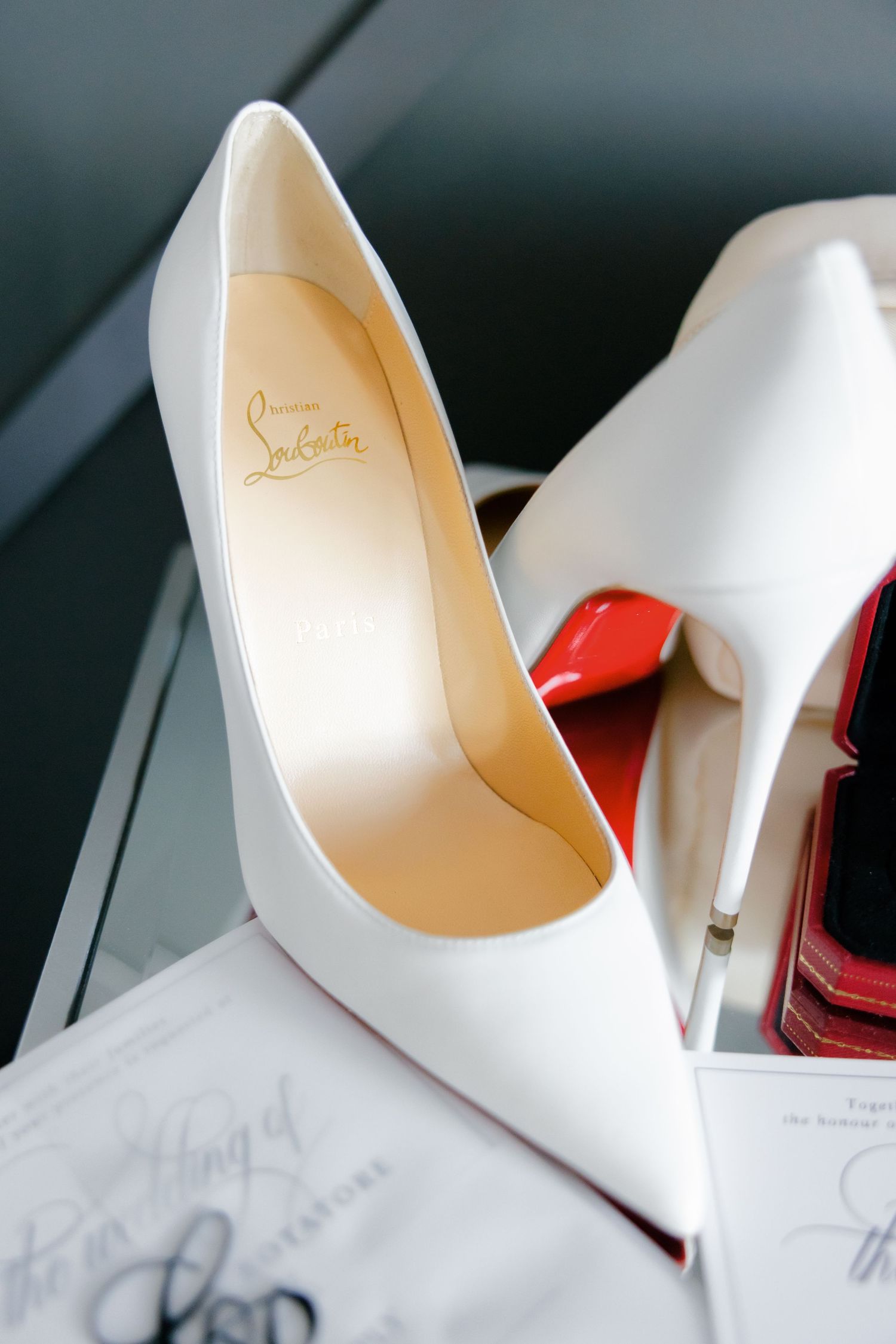 White Christian Louboutin stiletto heel shoe on reflective surface with wedding invitation.