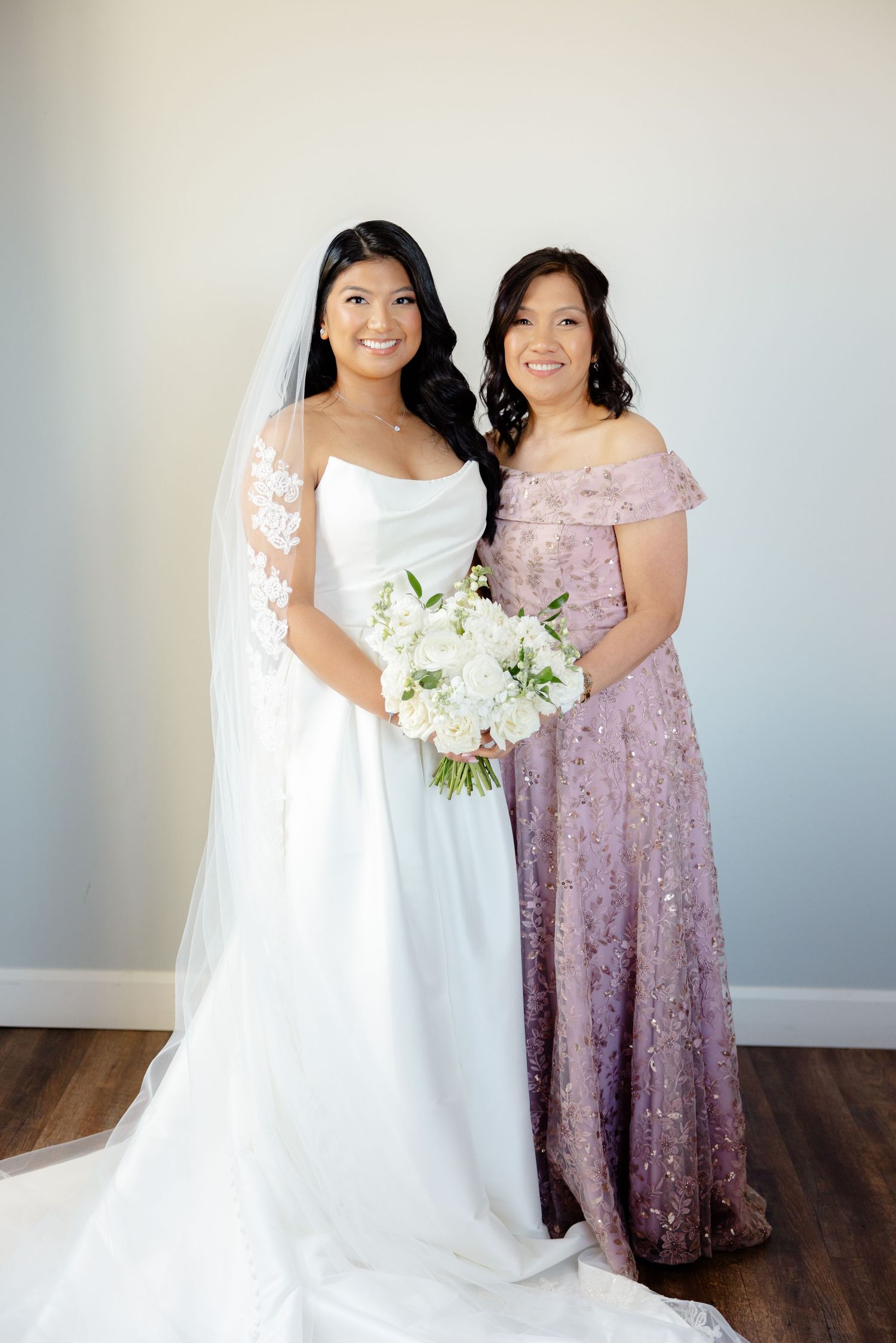 Bride in white gown and woman in lavender dress pose together with white floral bouquet.