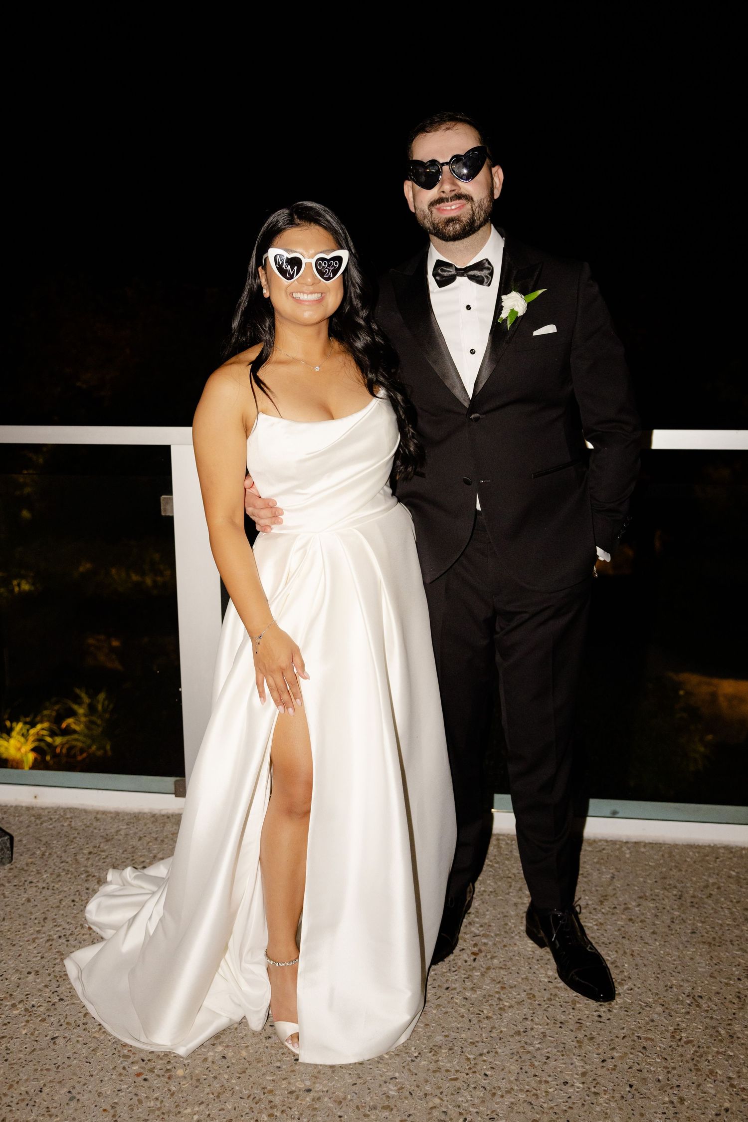 Stylish couple in wedding attire and sunglasses pose on outdoor balcony.