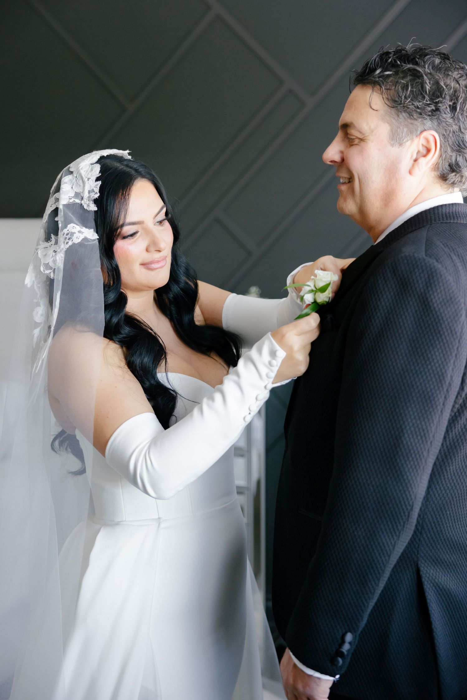 A bride in white adjusts a boutonniere on a dark suit during a wedding preparation moment.