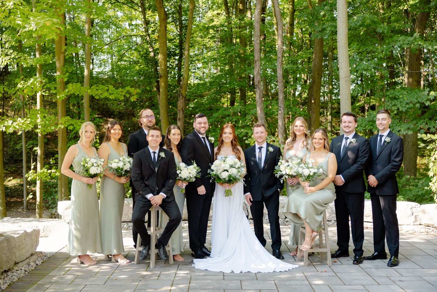 Wedding party poses on stone path in the woods with mix of sage green dresses and dark suits.
