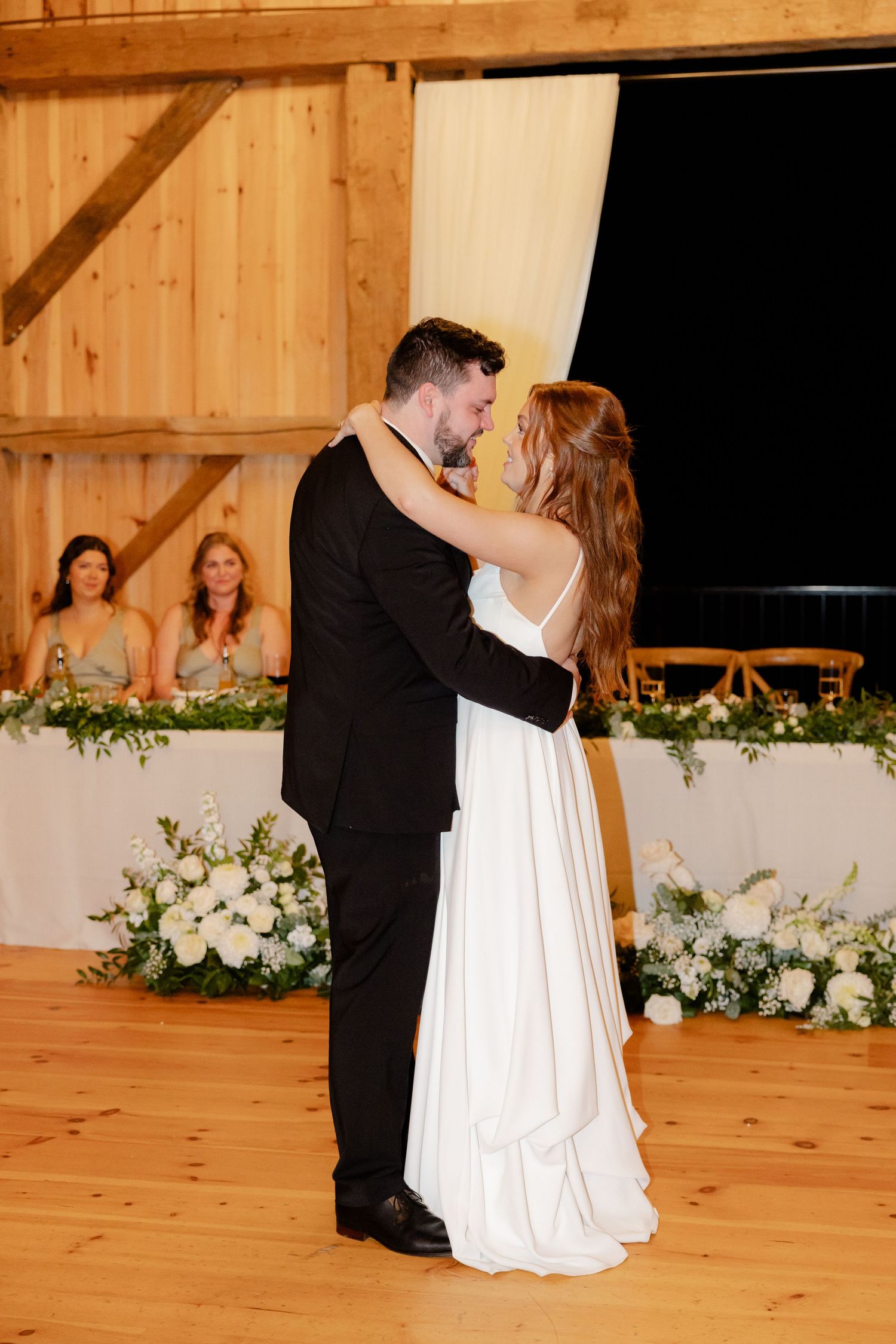 A couple dances in a rustic barn venue with bridesmaids and floral arrangements visible in background.