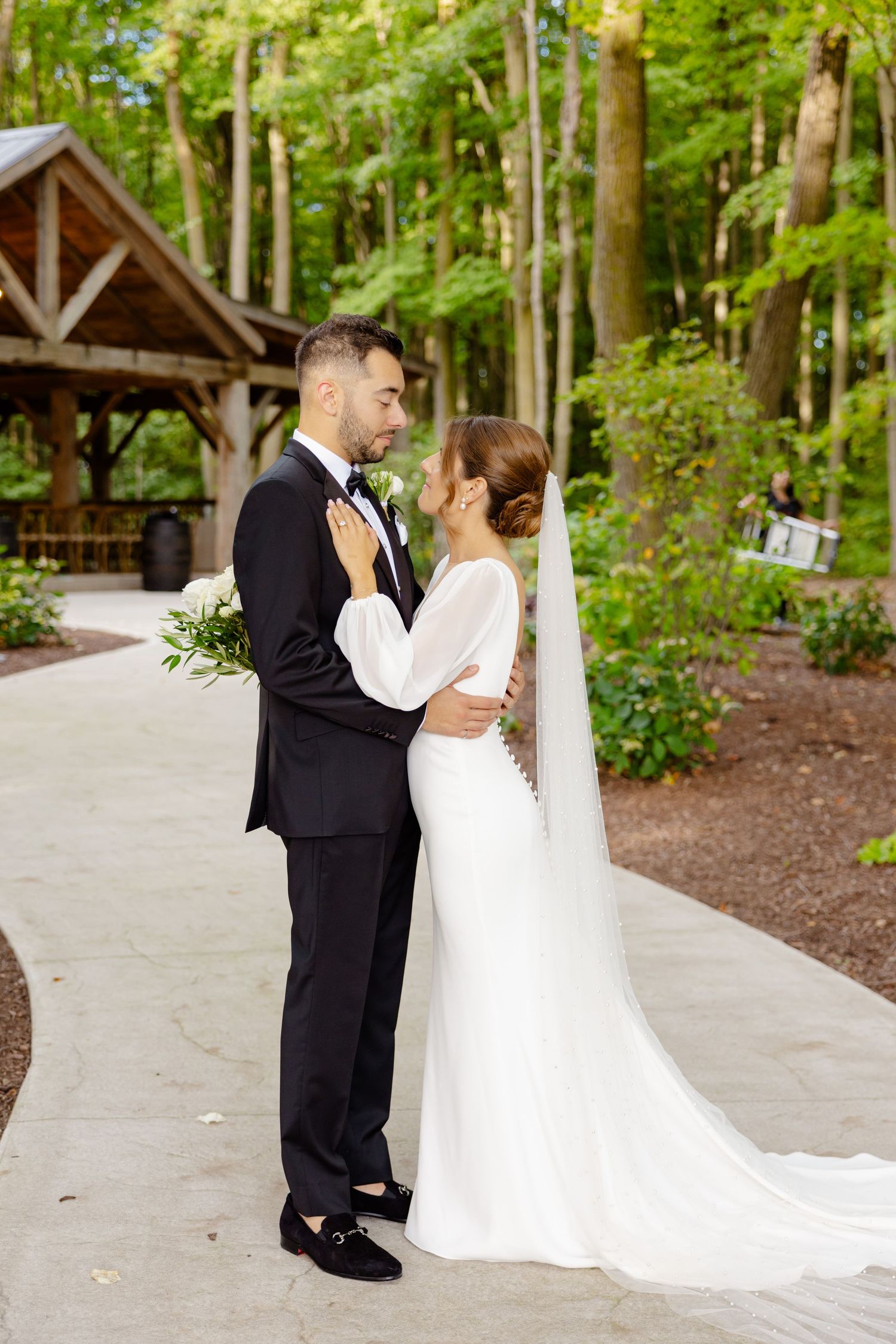 Intimate moment between newlyweds on concrete path with forest pavilion in background.