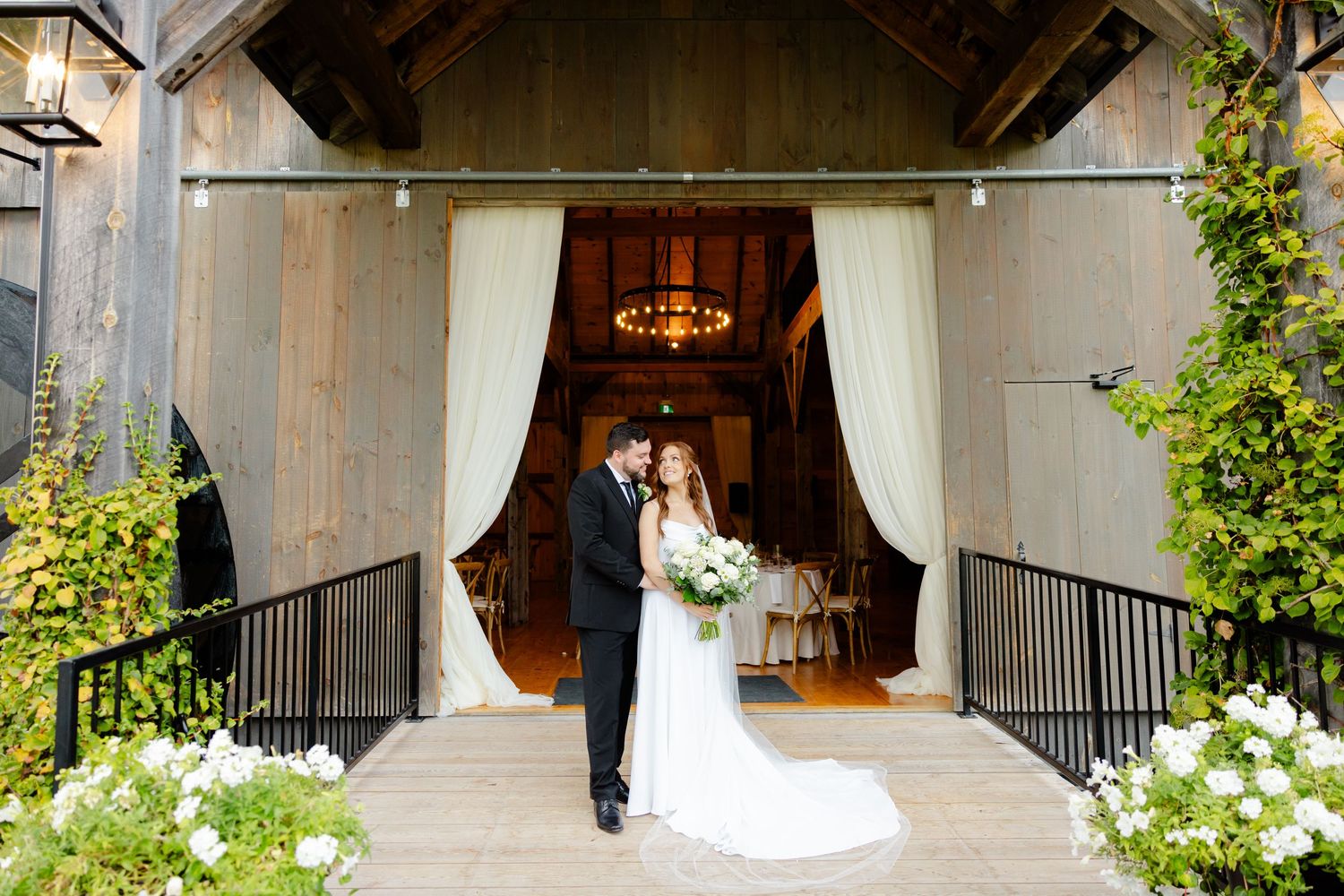 Wedding portrait at rustic barn entrance with white flowers and elegant drapery.