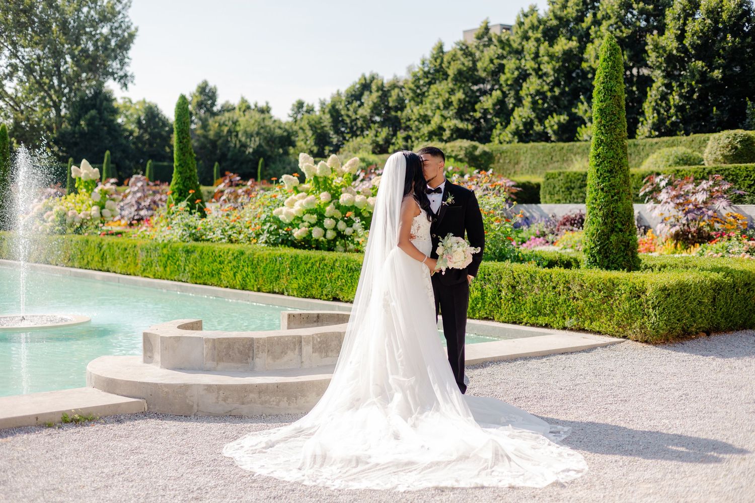 Newlyweds pose by an elegant fountain in a manicured garden with blooming flowers.