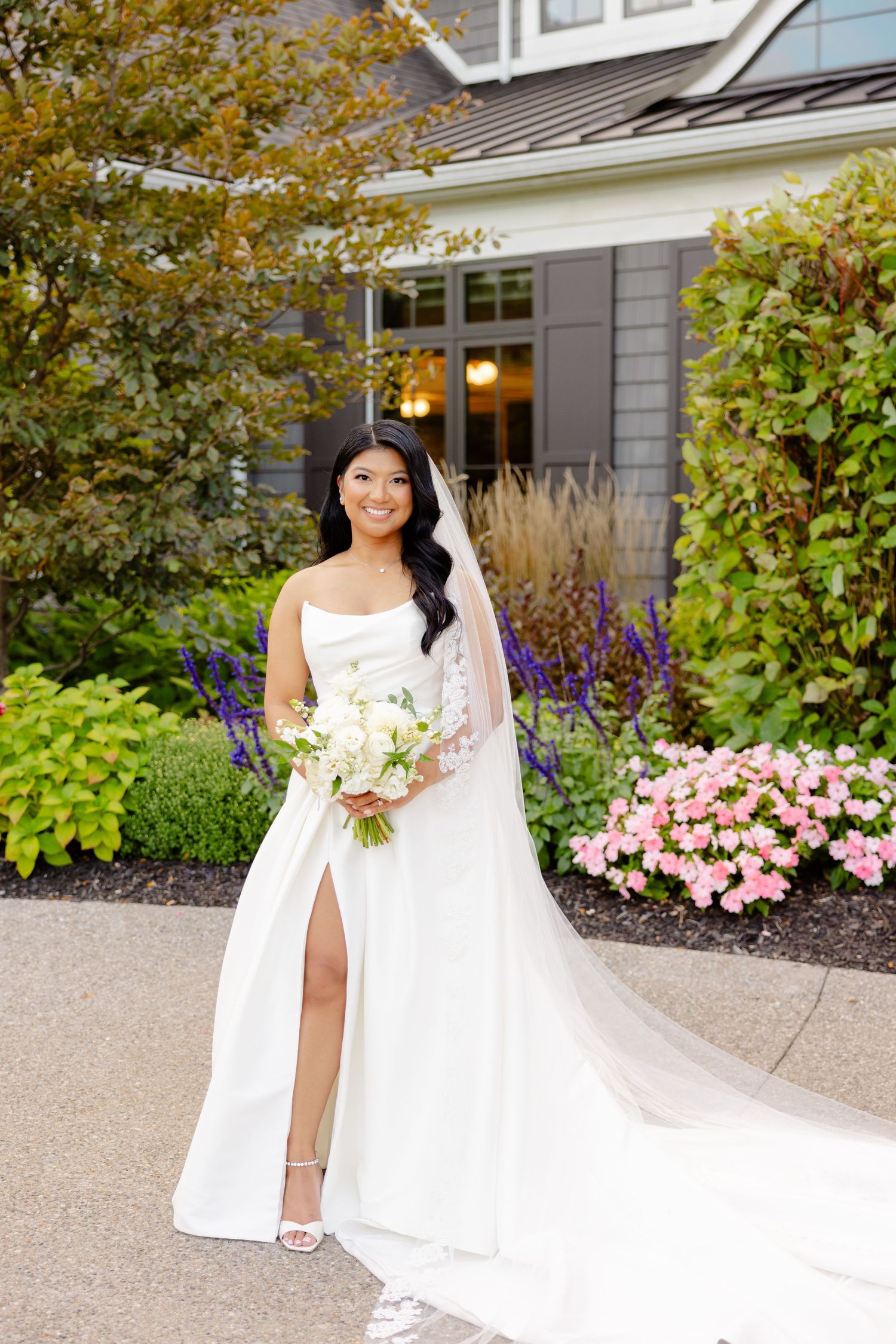 A stunning wedding dress with thigh slit and long train photographed in front of a grey house with beautiful landscaping.