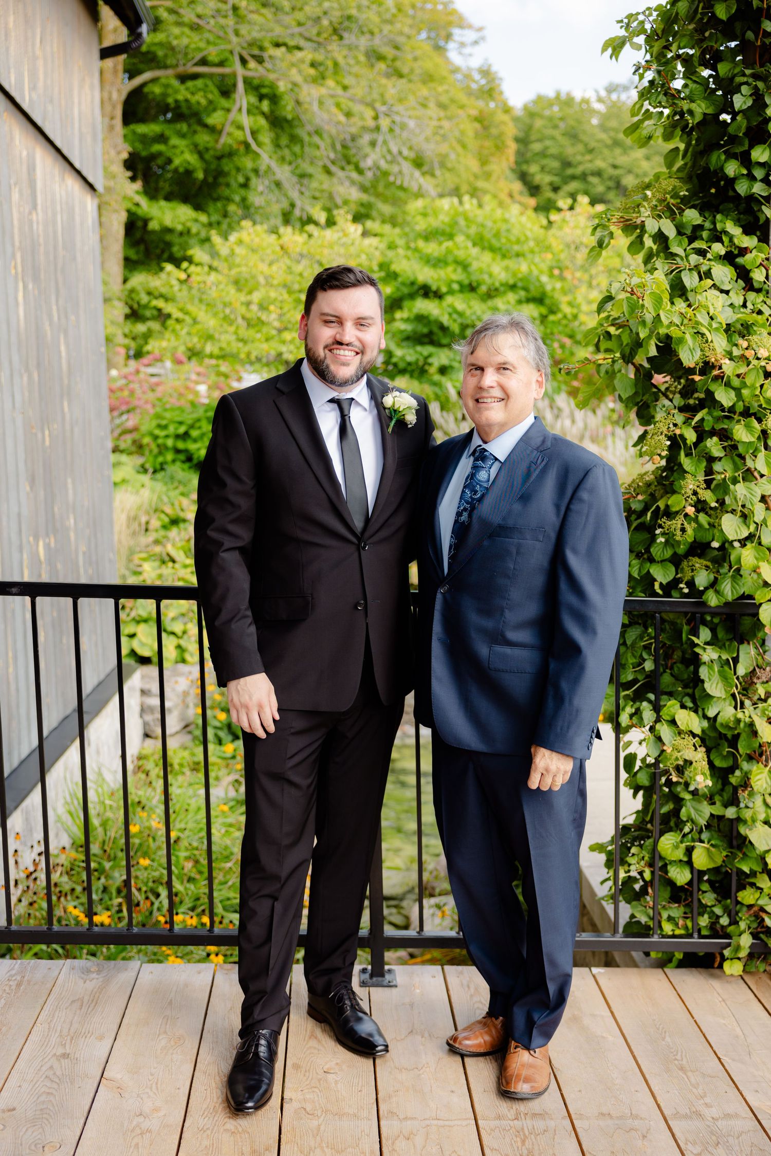 Two people in suits stand together on outdoor deck with green foliage backdrop.