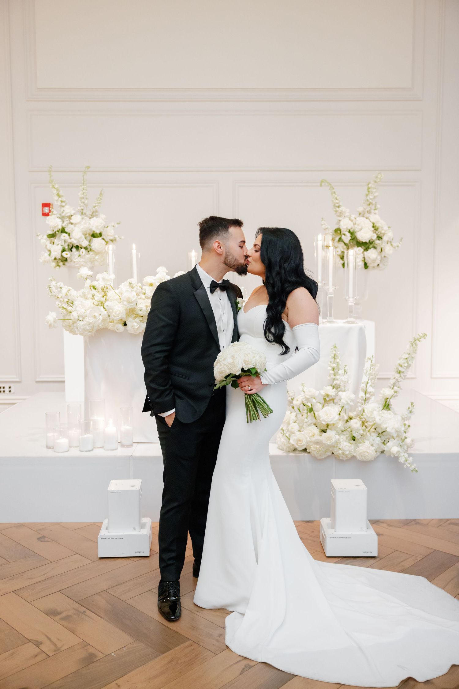 Wedding couple in classic attire embraces by white floral arrangements on herringbone floors.