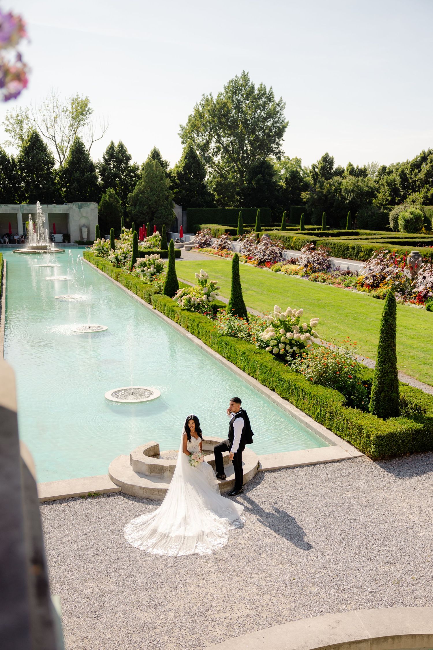 An elegant long pool with fountains surrounded by manicured gardens and cypress trees creates a romantic wedding setting.