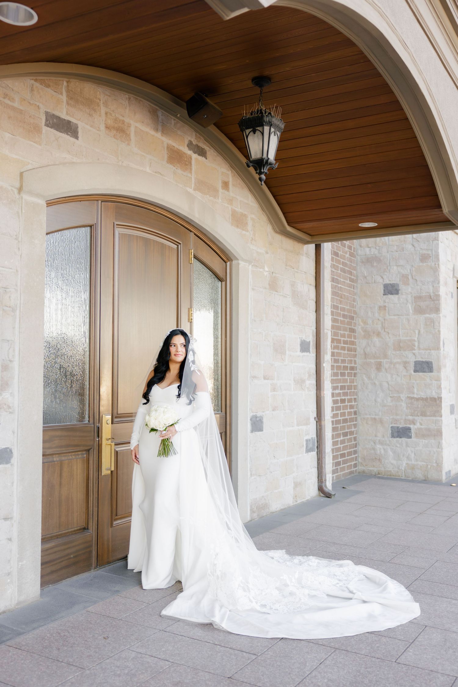 Bride in elegant white gown with cathedral veil poses in stone archway.