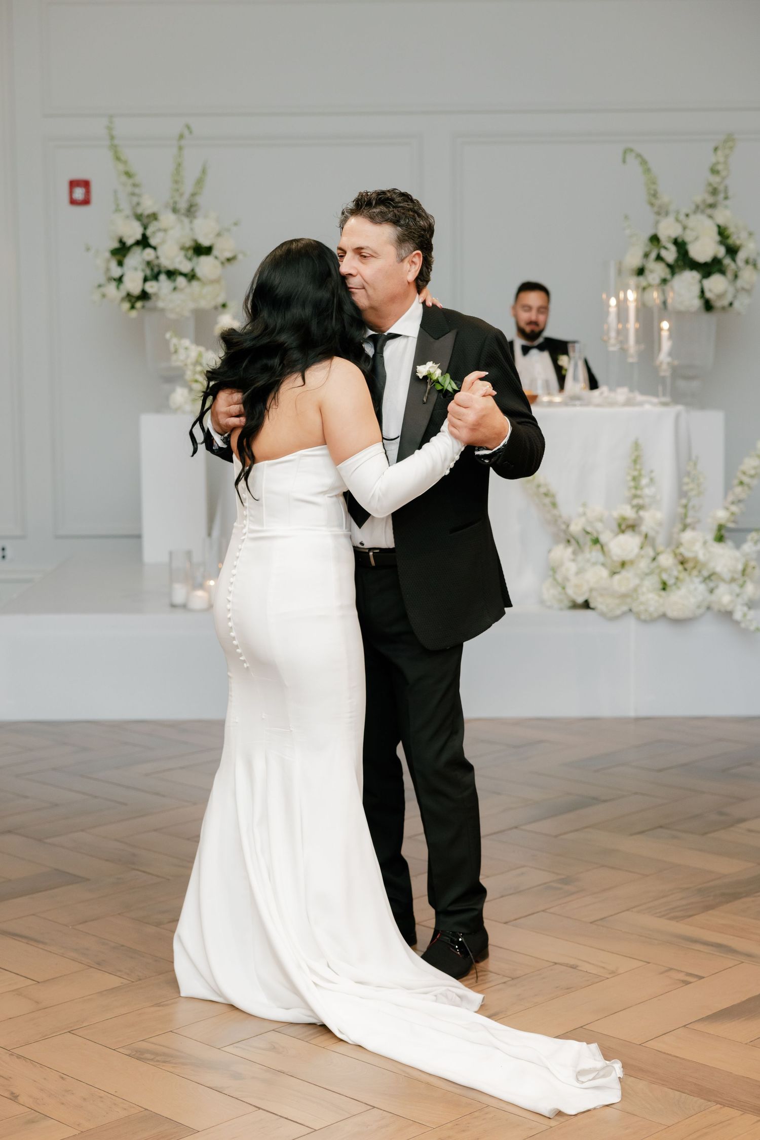 A couple dances on a herringbone wood floor surrounded by elegant white floral arrangements.