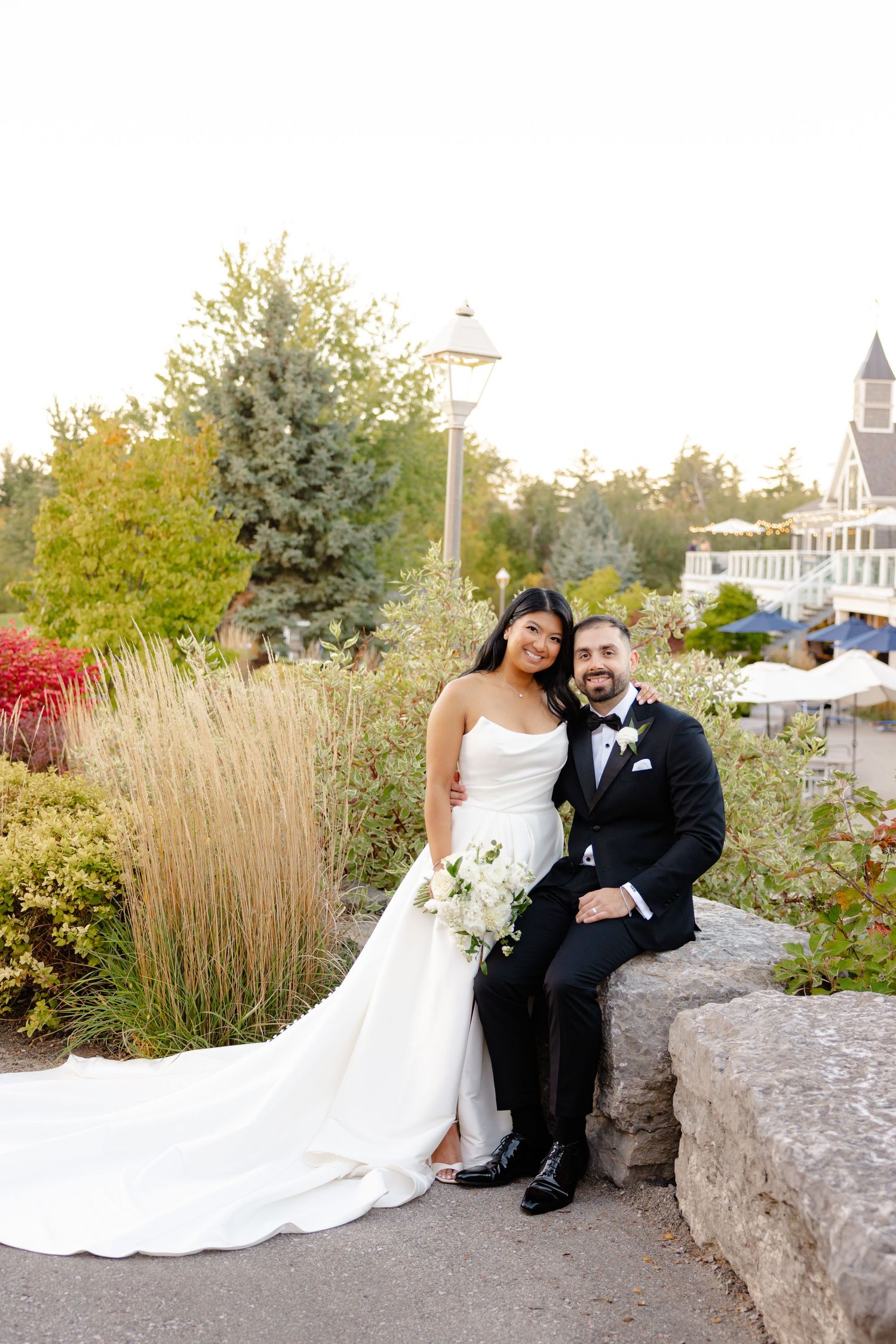A wedding couple poses near ornamental grasses and landscaping outside a beautiful venue.