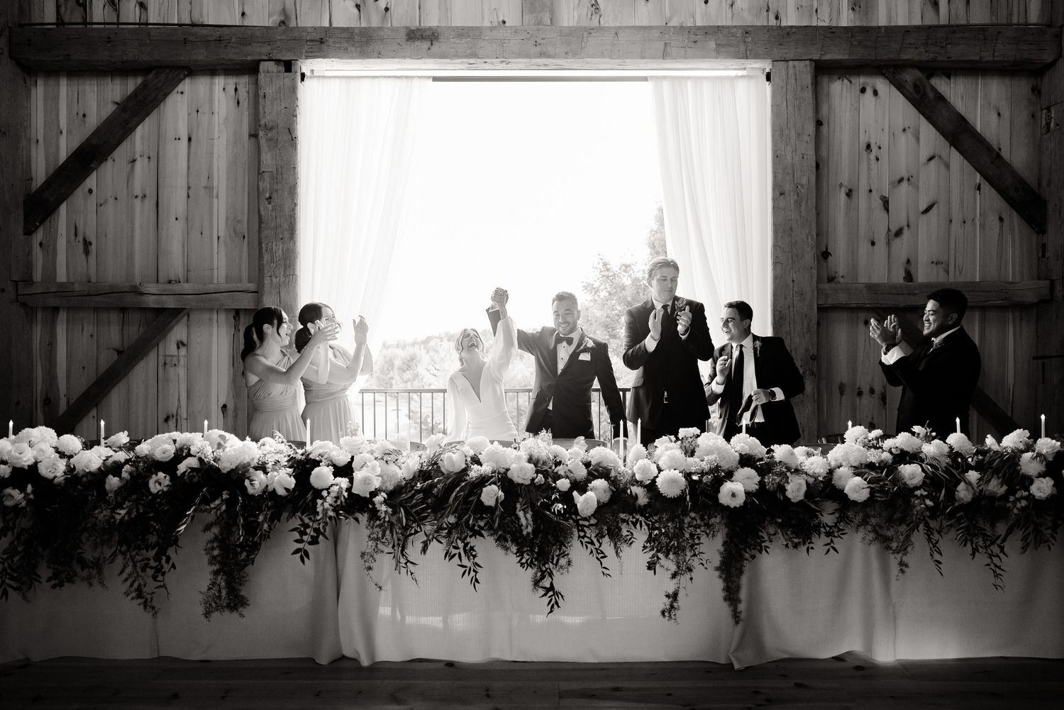 Wedding party lined up at head table decorated with cascading white floral arrangements in rustic barn venue.