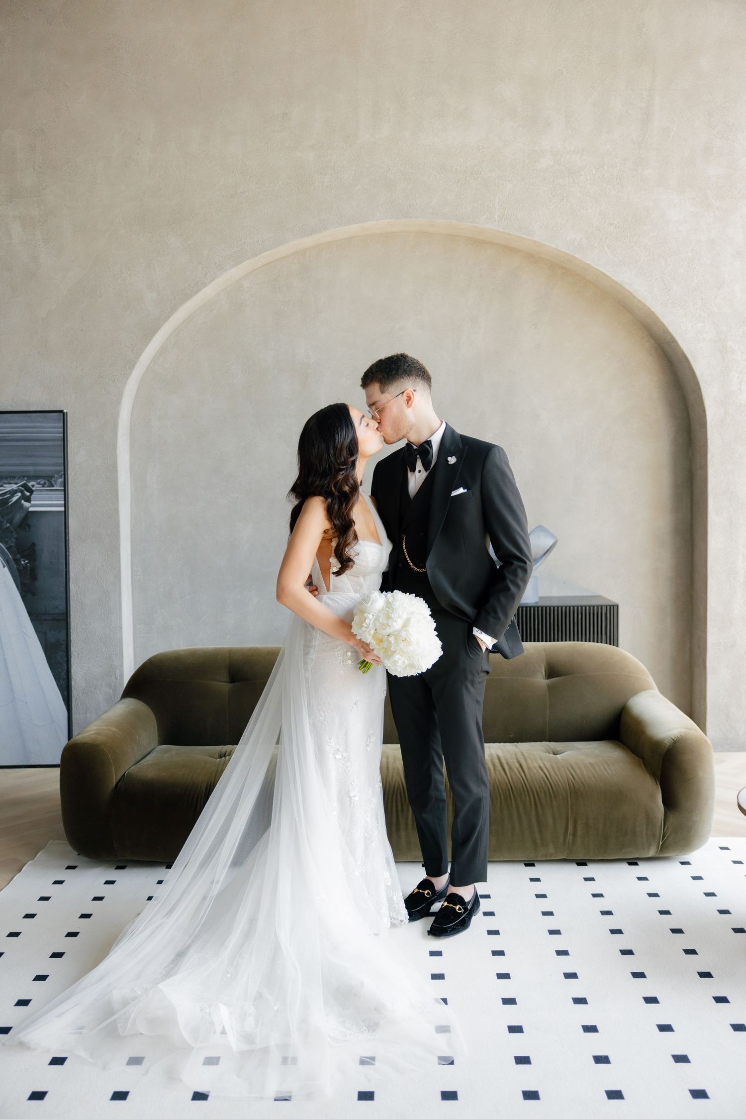 Elegant wedding couple shares tender moment against arched wall and olive couch.