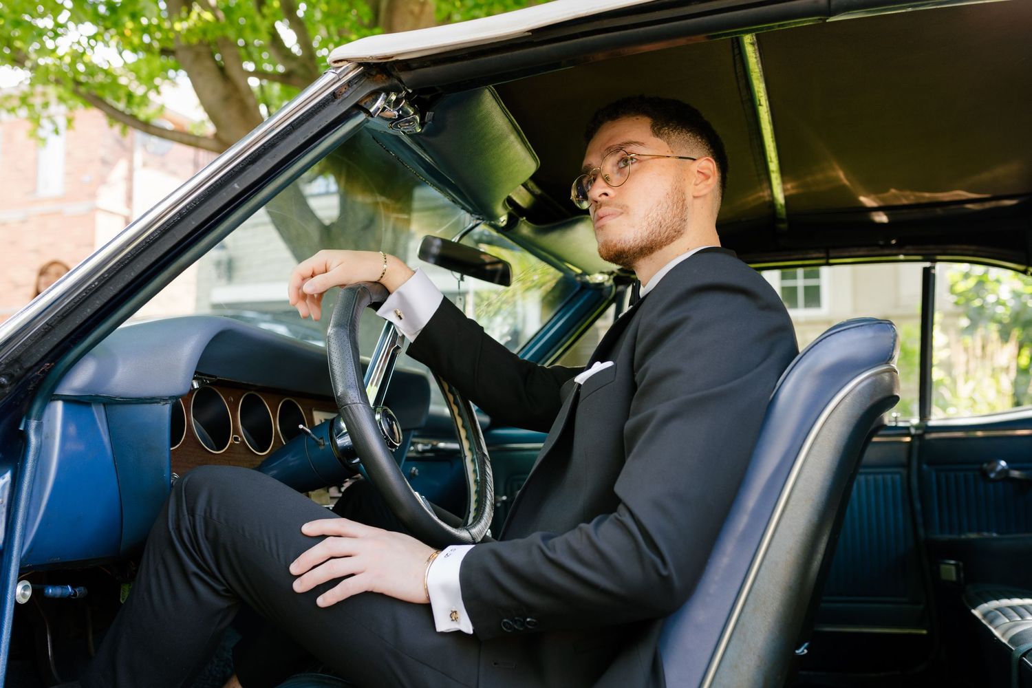 Person in formal suit sitting in driver's seat of vintage car interior.