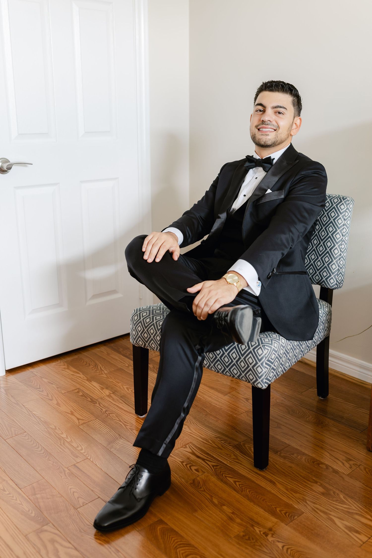 Formal portrait of person in tuxedo seated in patterned chair.