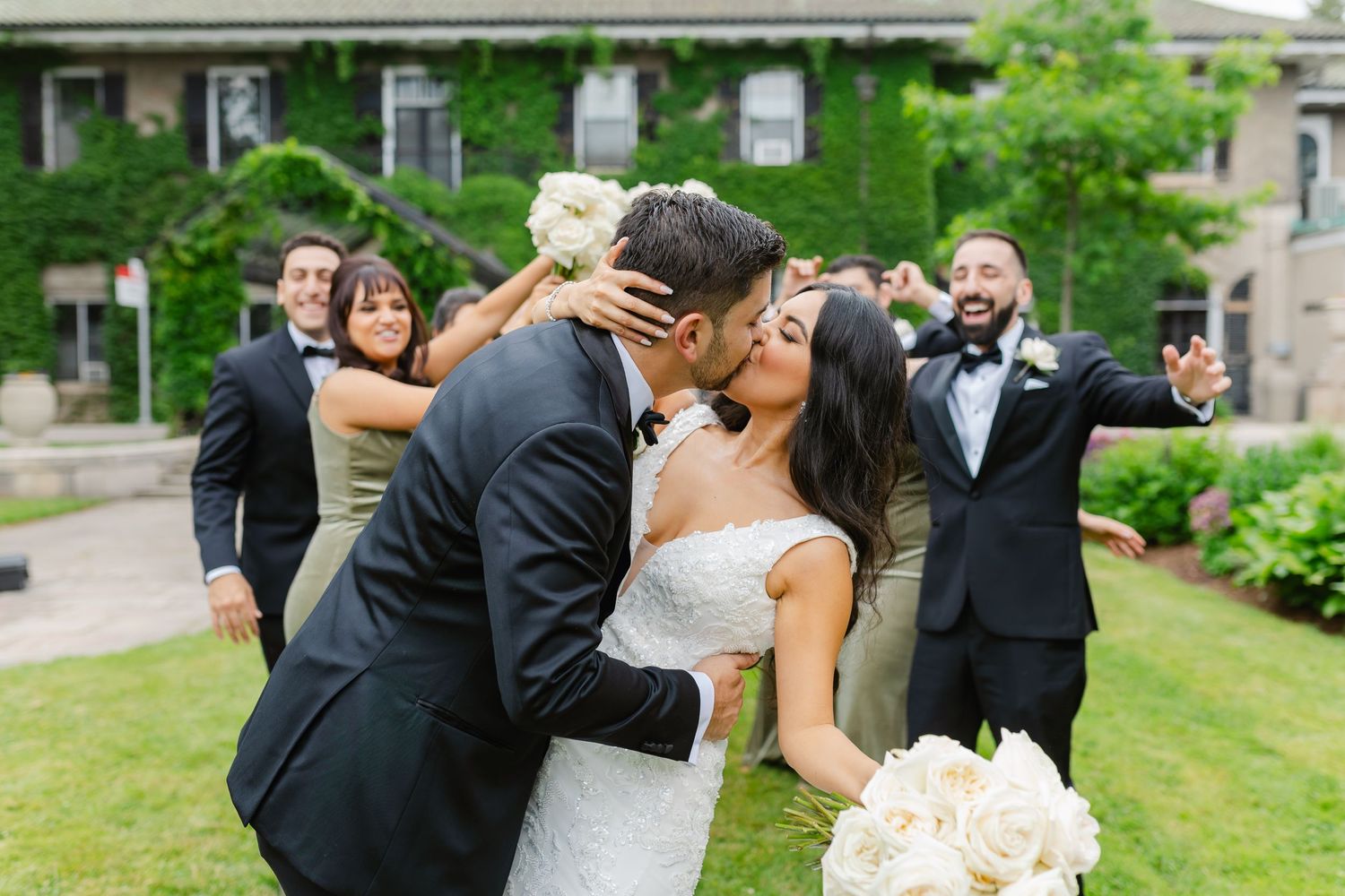 Wedding couple shares romantic moment surrounded by celebrating bridal party on mansion lawn.