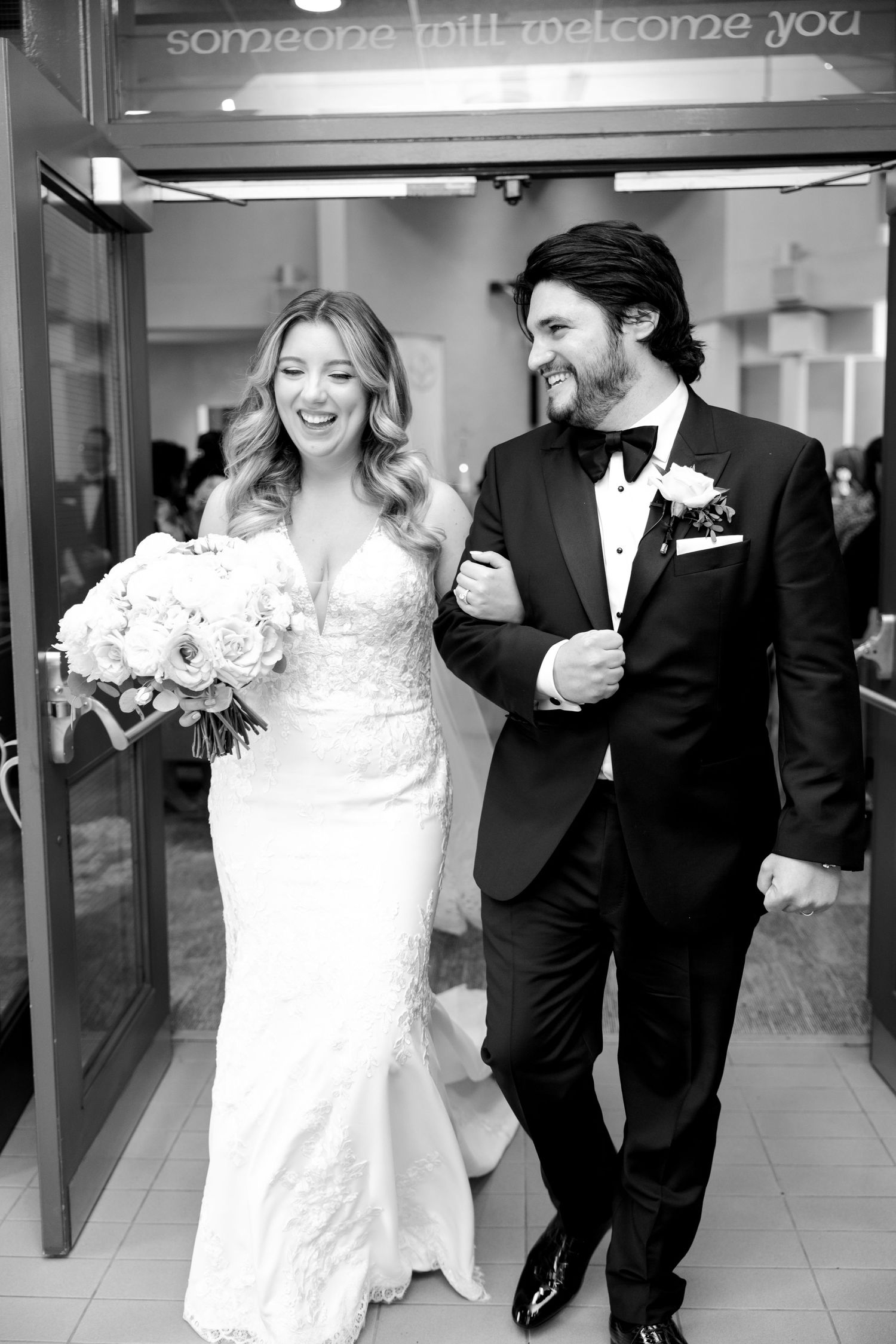 Wedding couple laughing as they exit the church under a welcome sign.