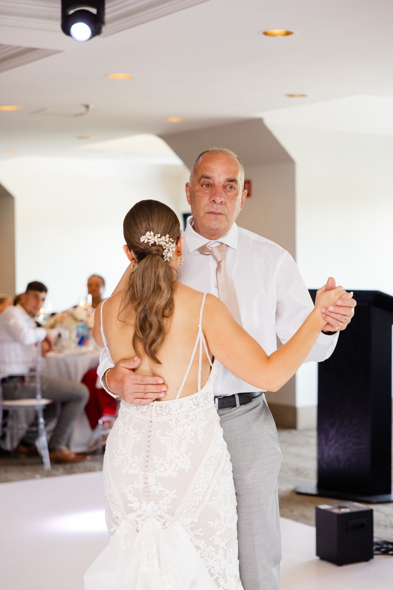 Couple dances in an elegant white ballroom setting with guests seated in the background.