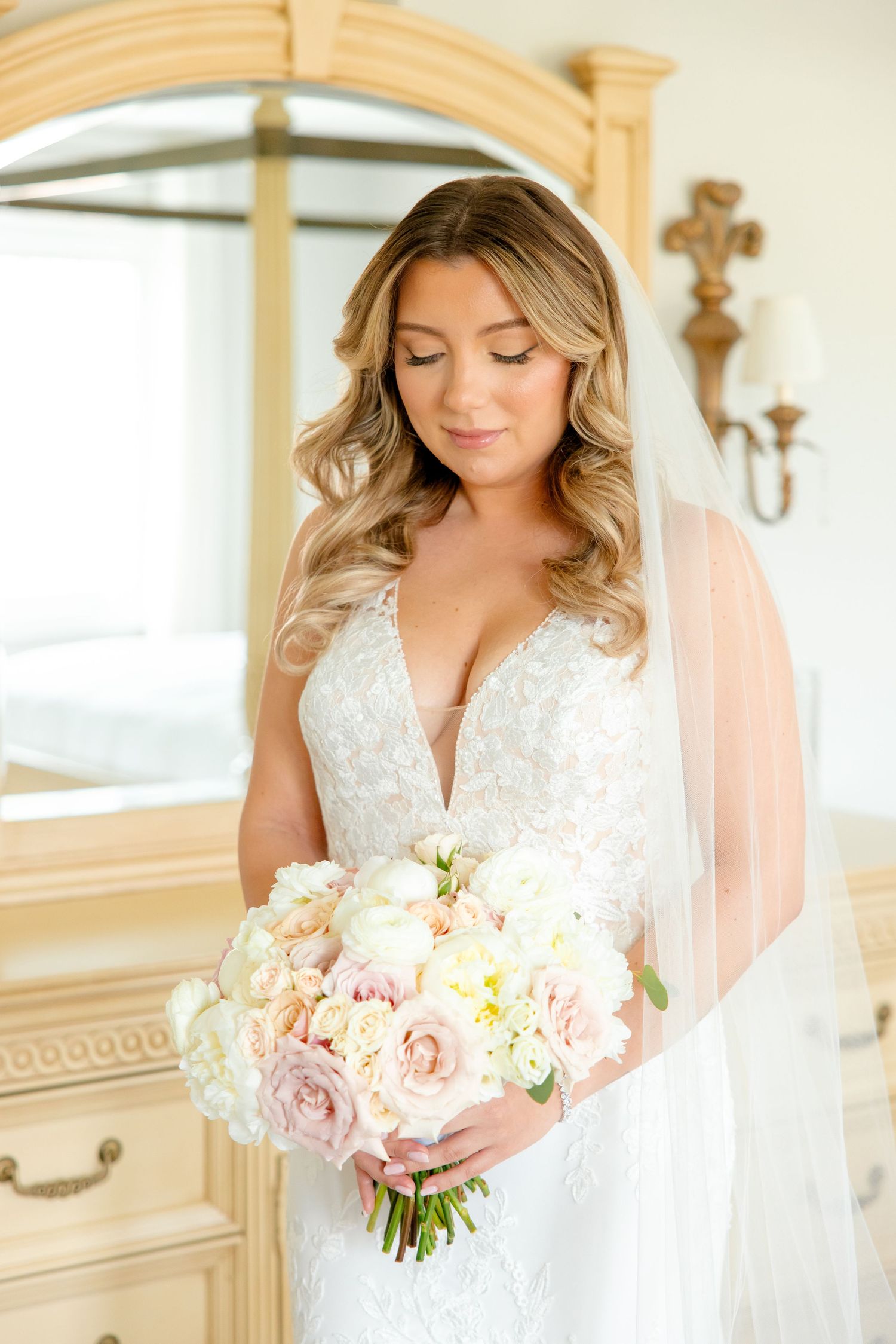 Romantic bridal portrait showcasing detailed lace wedding gown and pastel bouquet against light wooden furniture.