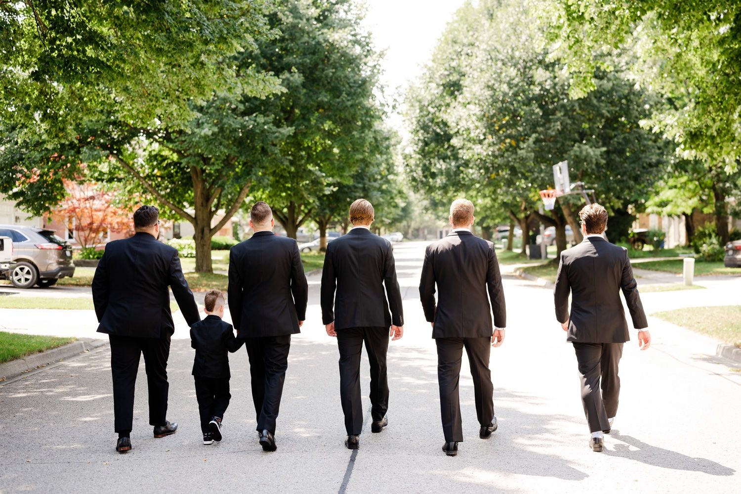 Wedding party in black suits walking down a tree-lined path.