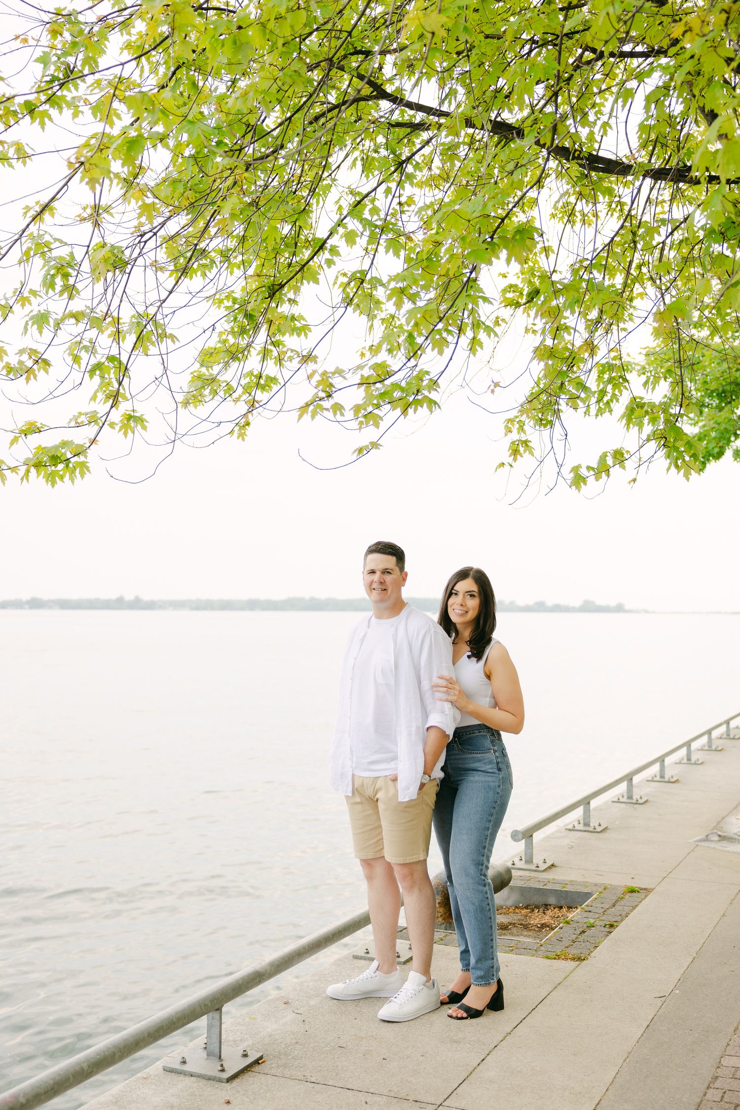 A couple stands together by the waterfront under overhanging green tree branches.