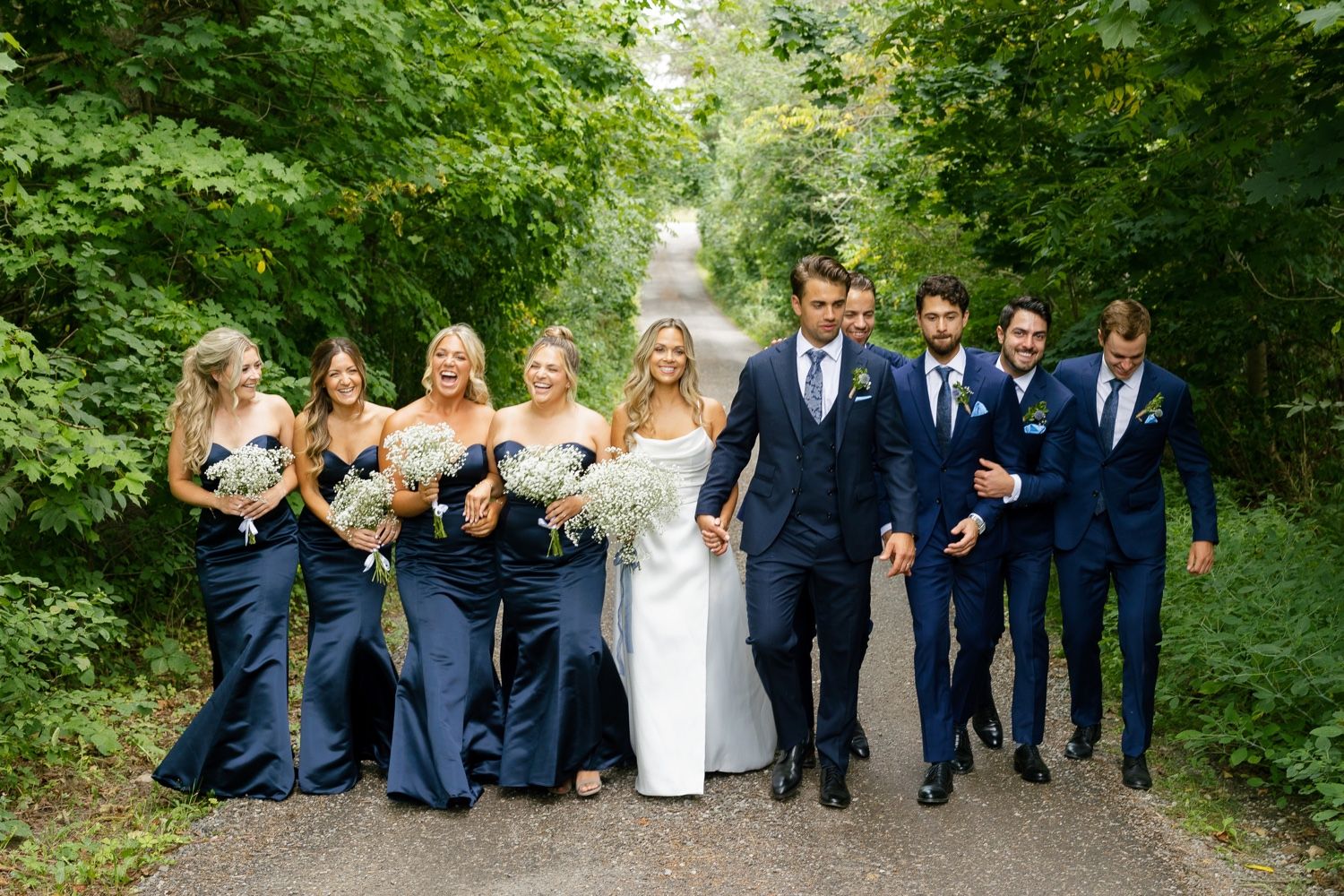Wedding party walking together on tree-lined path with bridesmaids in navy dresses.