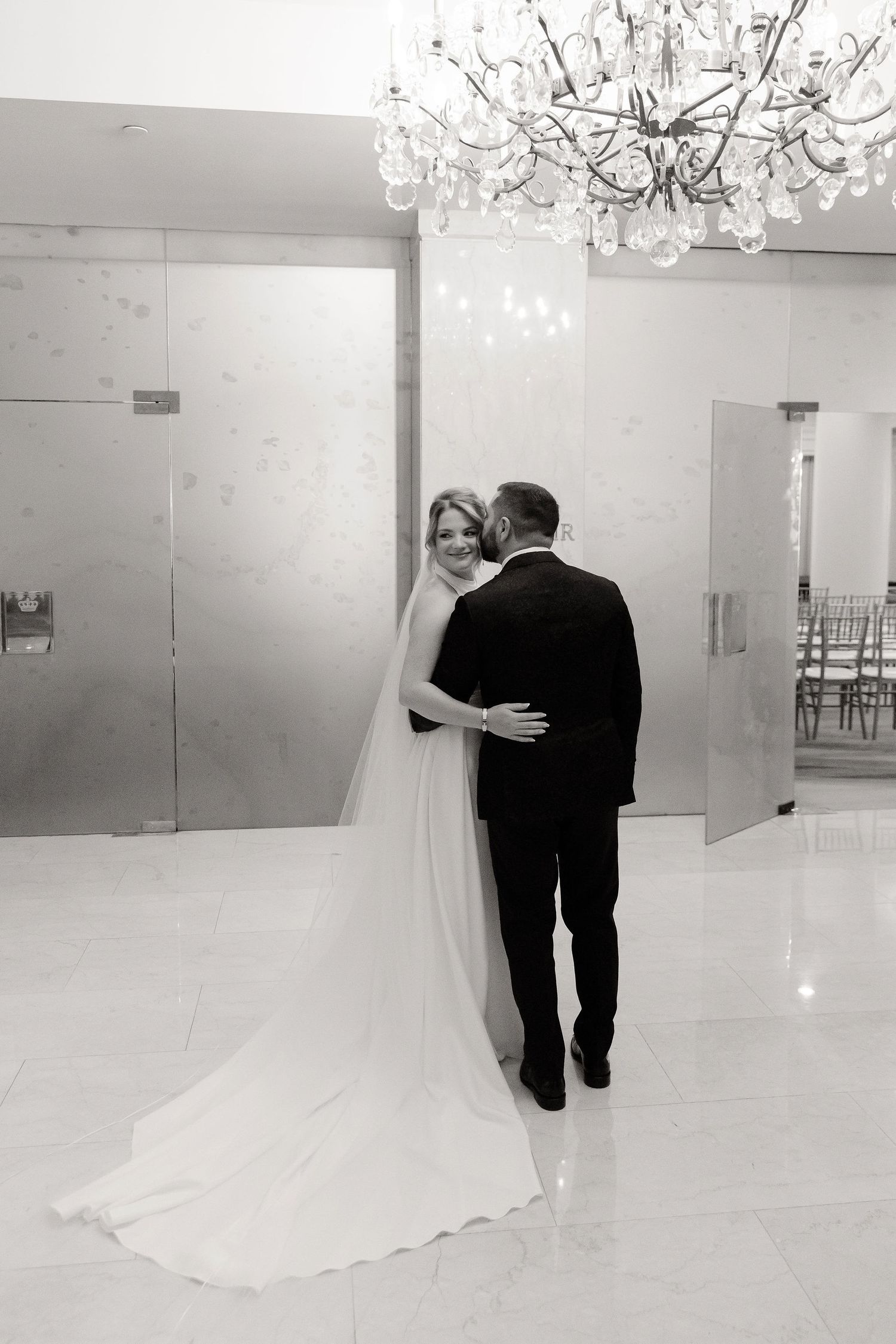 Romantic wedding moment under a crystal chandelier in an elegant white room.