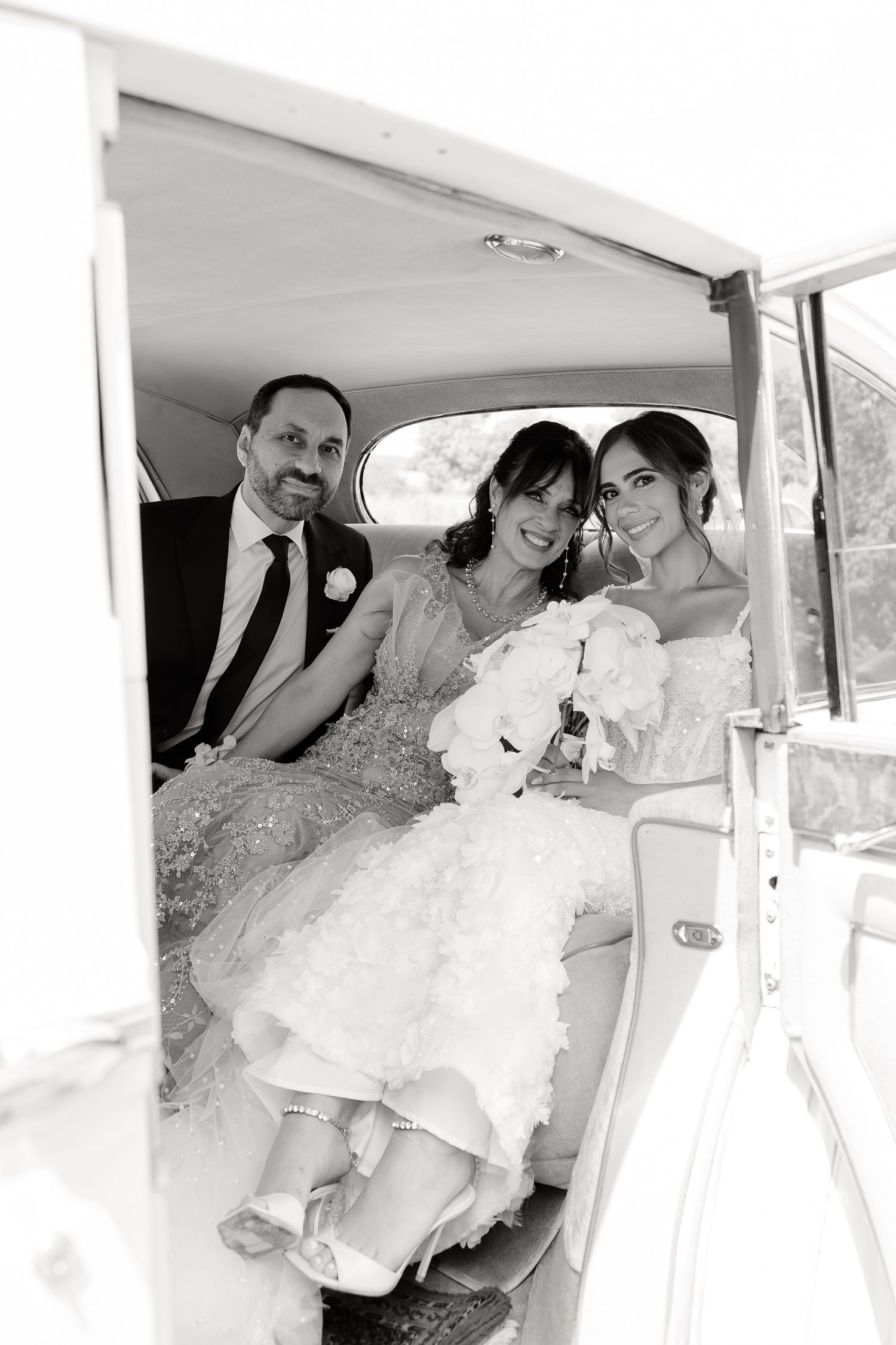 Black and white photo of wedding party inside vintage car interior with white flowers and formal attire.