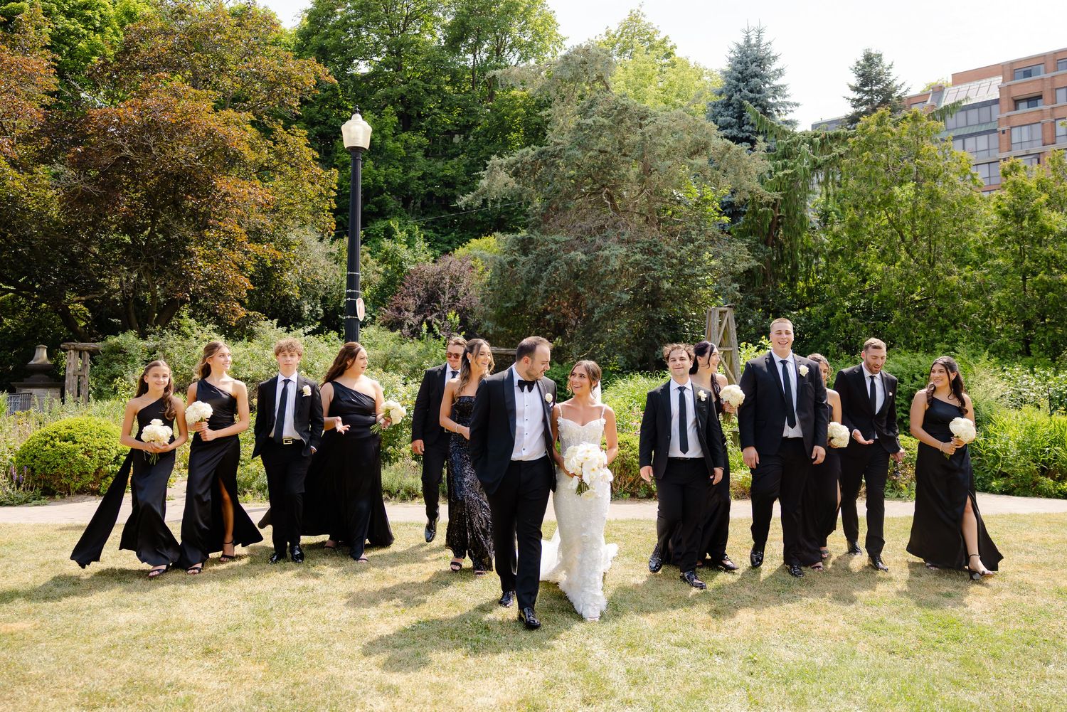 A wedding party dressed in black and white formal attire poses together outdoors on a grassy hillside in summer.