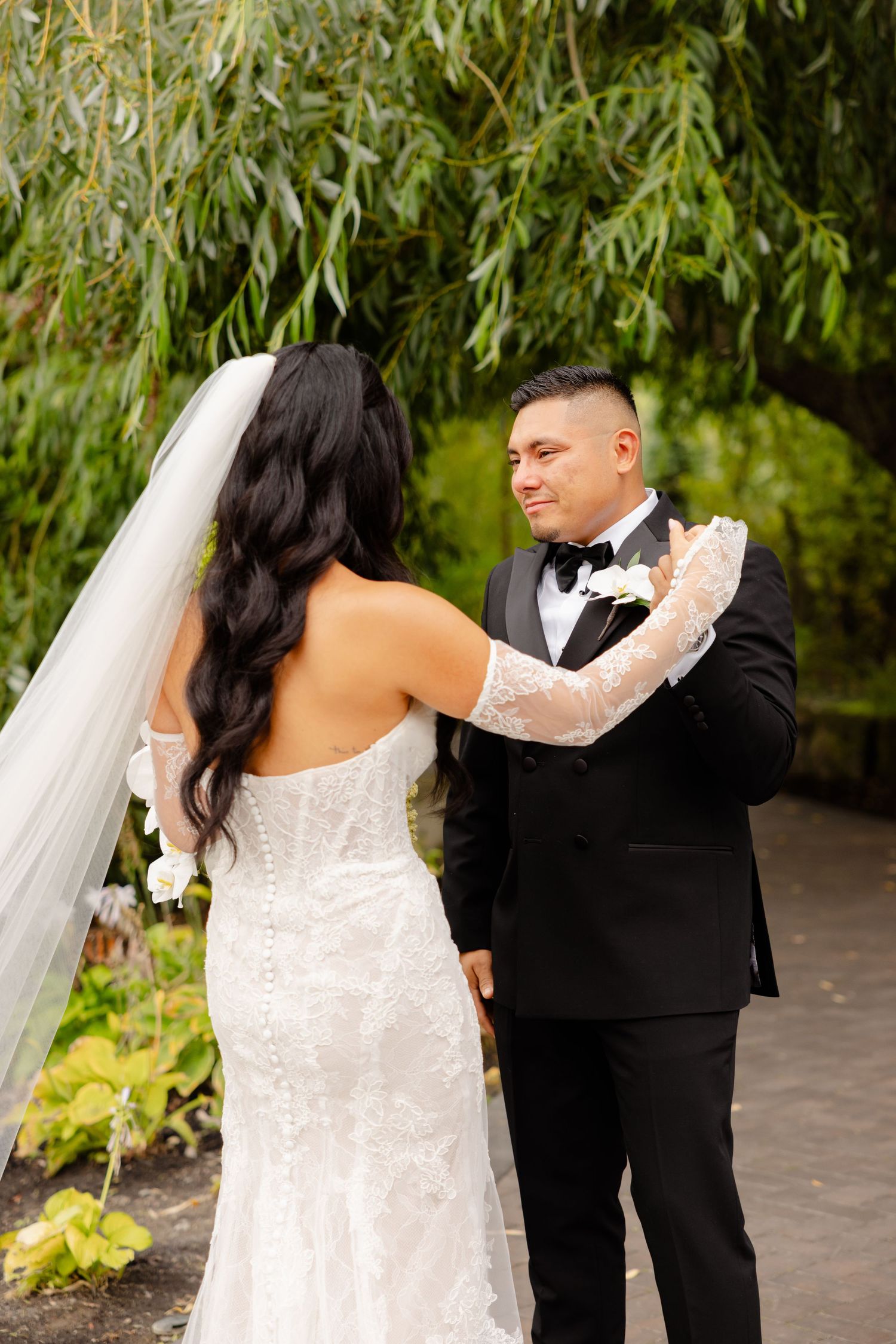 A couple in elegant wedding attire dance beneath willow tree branches in a romantic garden setting.