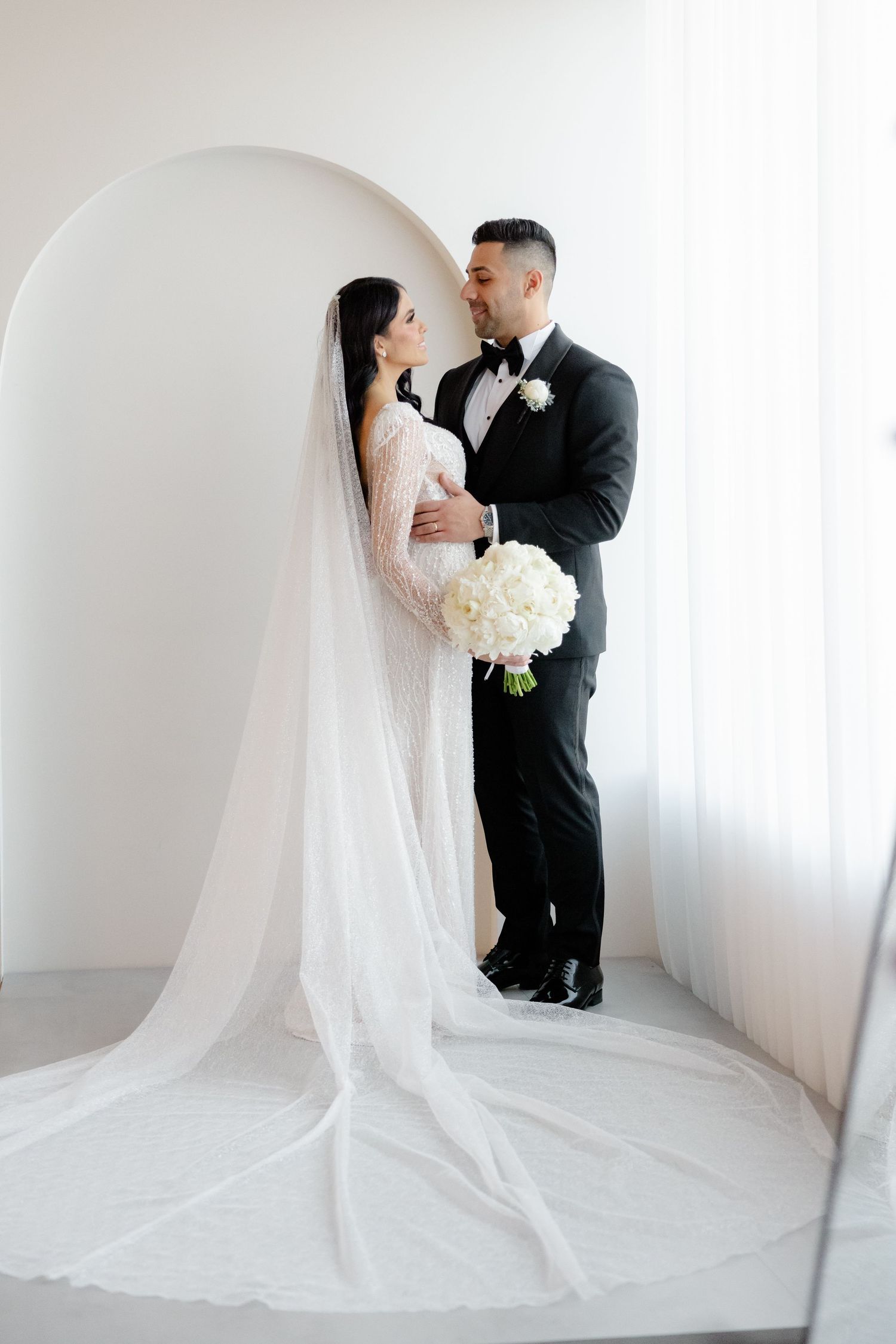 Wedding couple shares tender moment in minimalist white setting with cathedral veil.