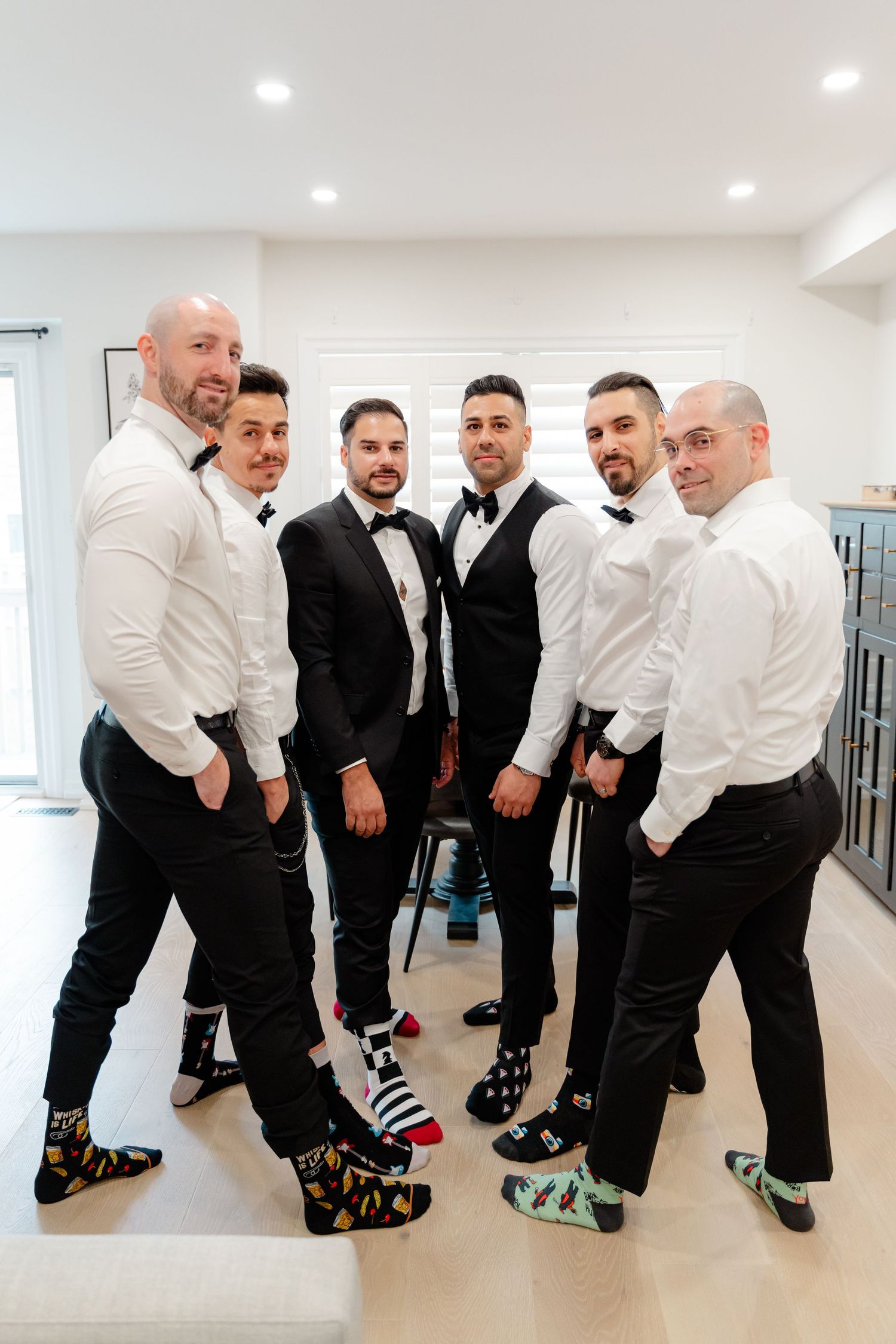 Group of groomsmen in formal wear posing to show off their colorful socks in a modern room.