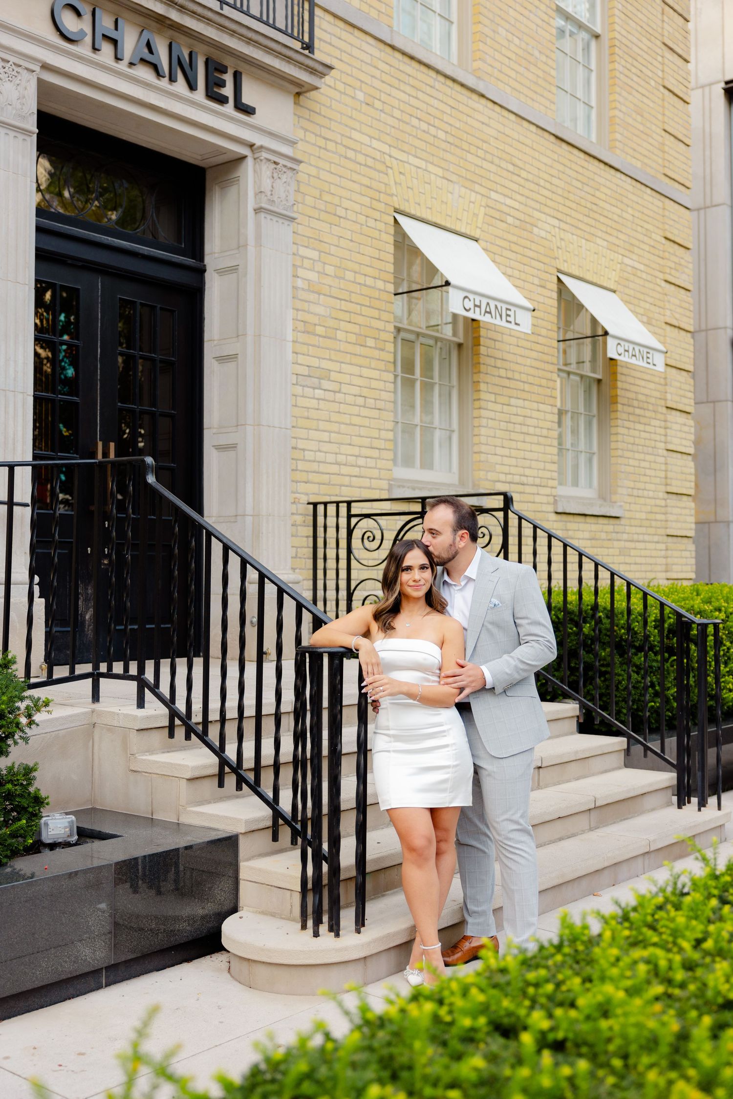 Fashionable pair stands together on steps of luxury Chanel store with ornate iron railings.
