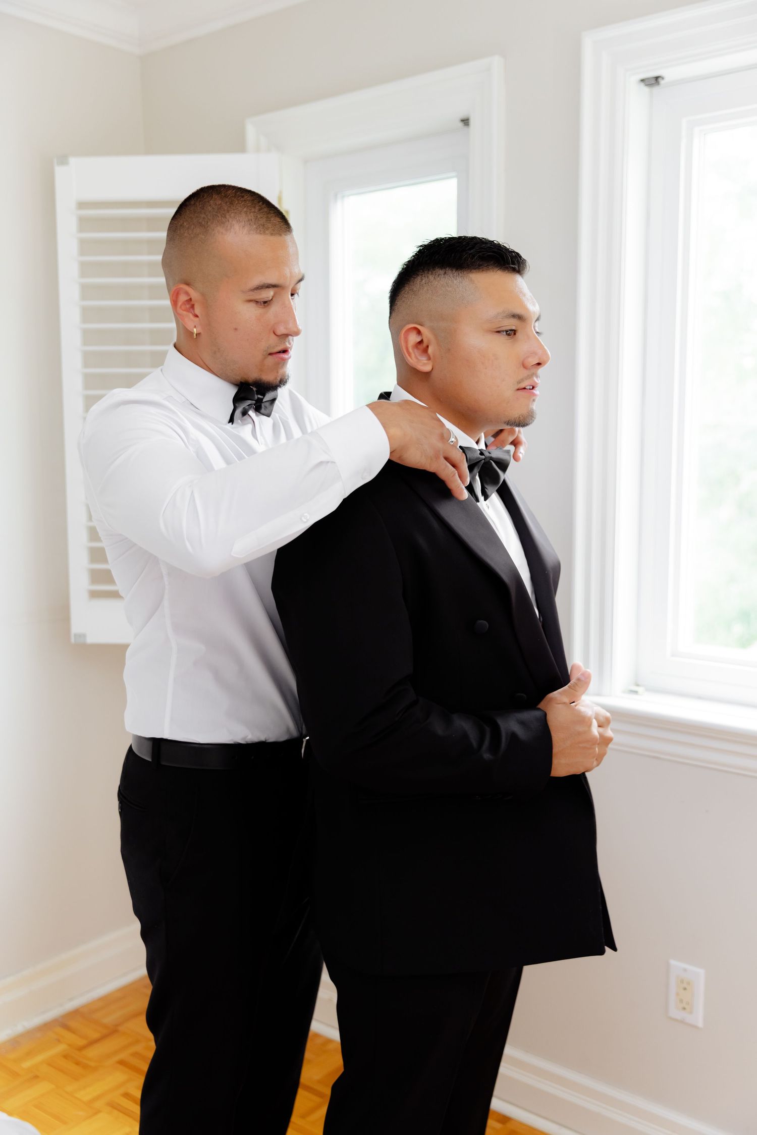 A groomsman helps another adjust his tie before a wedding ceremony in a bright white room with hardwood floors.
