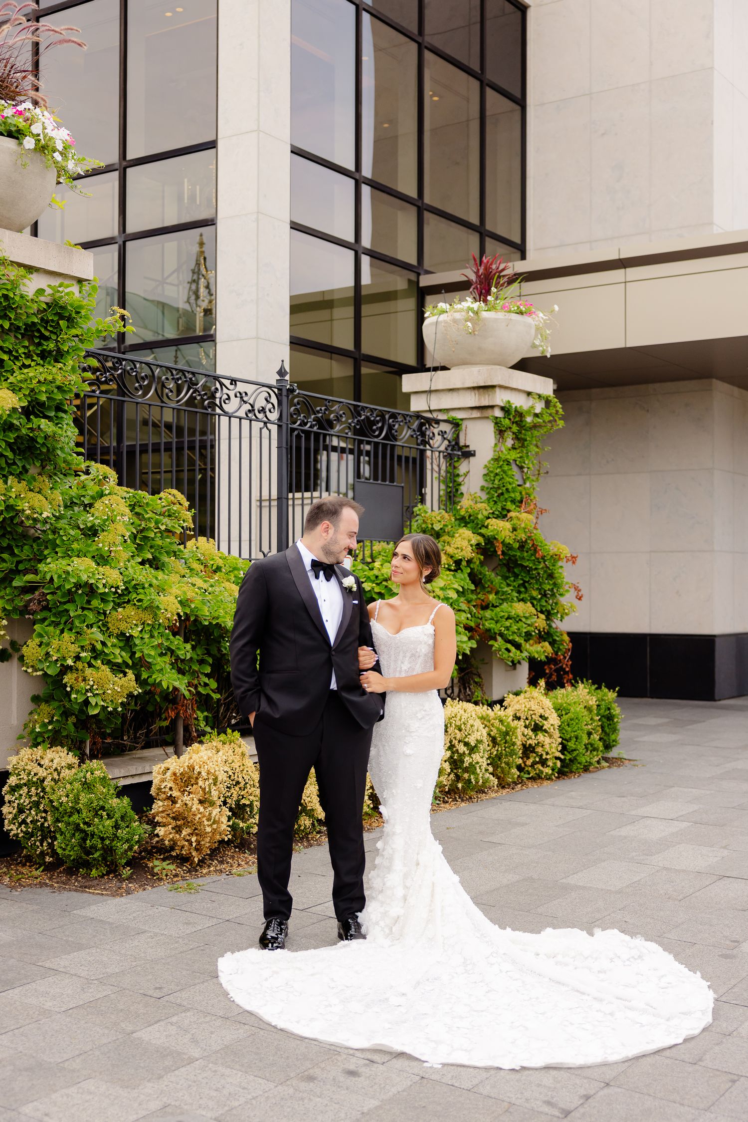 A happy couple in formal wedding attire share an intimate moment on a patio with greenery and columns.